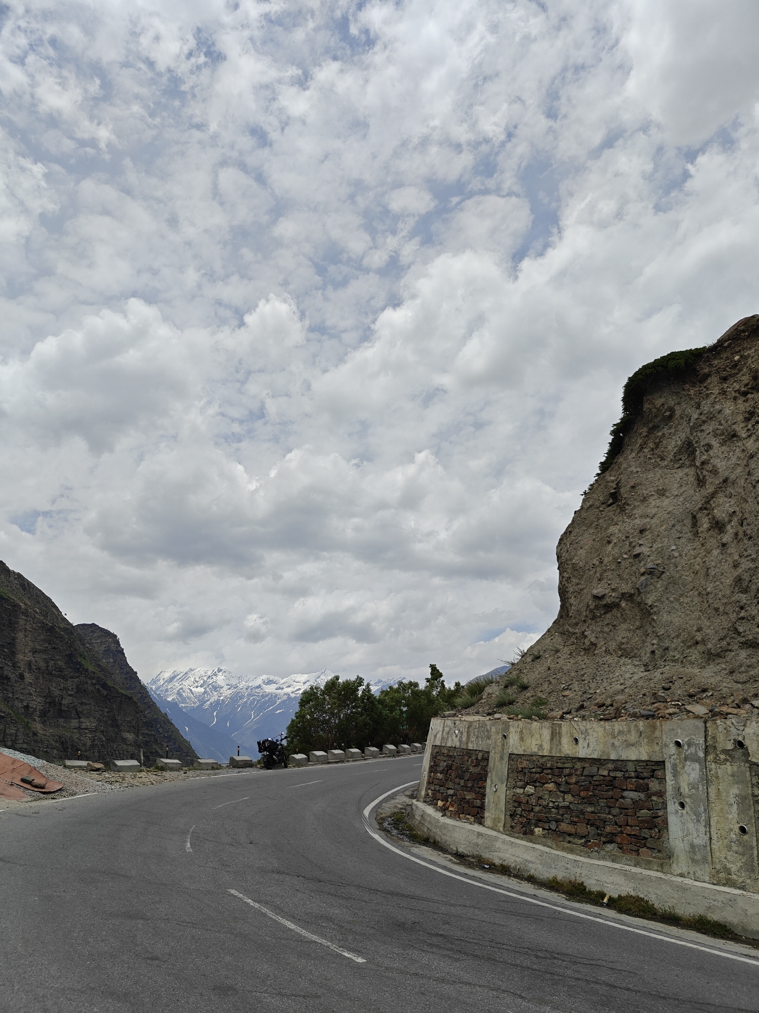 The image shows a winding asphalt road in a mountainous region of India. The road curves to the right, with a steep, rocky embankment on the right side and a dark, rocky mountain slope on the left. In the background, snow-capped mountains are visible under a cloudy sky. A motorcycle is parked on the side of the road on the right. The embankment on the right has a retaining wall constructed with concrete and stone. The environment appears to be a remote and rugged landscape, likely a mountain pass.