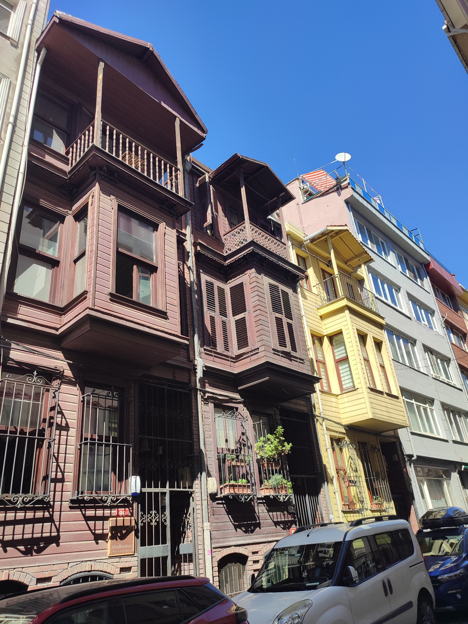 A street scene in Kadıköy, Turkey, features a row of buildings with varying architectural styles under a clear blue sky. The leftmost building is a traditional wooden structure with a protruding bay window and an upper-level balcony, both adorned with intricate wooden latticework. The building to its right is also wooden, featuring a covered balcony with shutters and similar bay window elements. Further down the street, the architecture shifts to more modern styles, with a pale pink building topped by a satellite dish and an awning, followed by a bright yellow building with a small balcony, and then a grey multi-story building with numerous windows. The lower portion of the image shows the street level, with several parked vehicles, including a white van and a dark blue SUV. A sign for "Khalkedon Film & Animation" is visible on the wooden building. The overall impression is a blend of historical and contemporary urban design in a Turkish city.