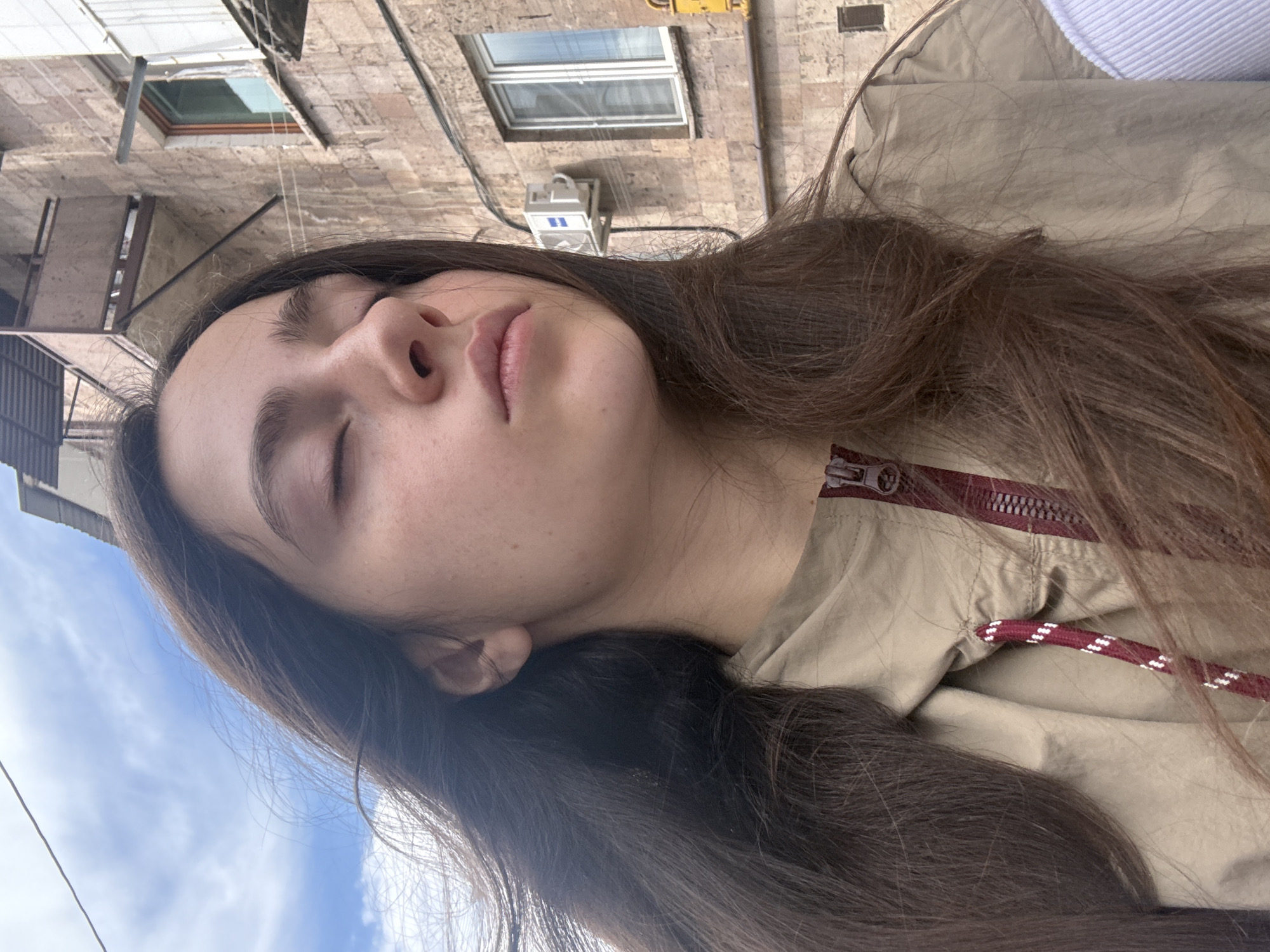 A photograph taken in Yerevan, Armenia shows a young woman with long, dark brown hair, closed eyes, and a fair complexion wearing a beige jacket with a maroon zipper and drawstring. The background displays the architecture of Yerevan, including a building with tan brick and multiple windows. The sky with light blue and white clouds is also visible.