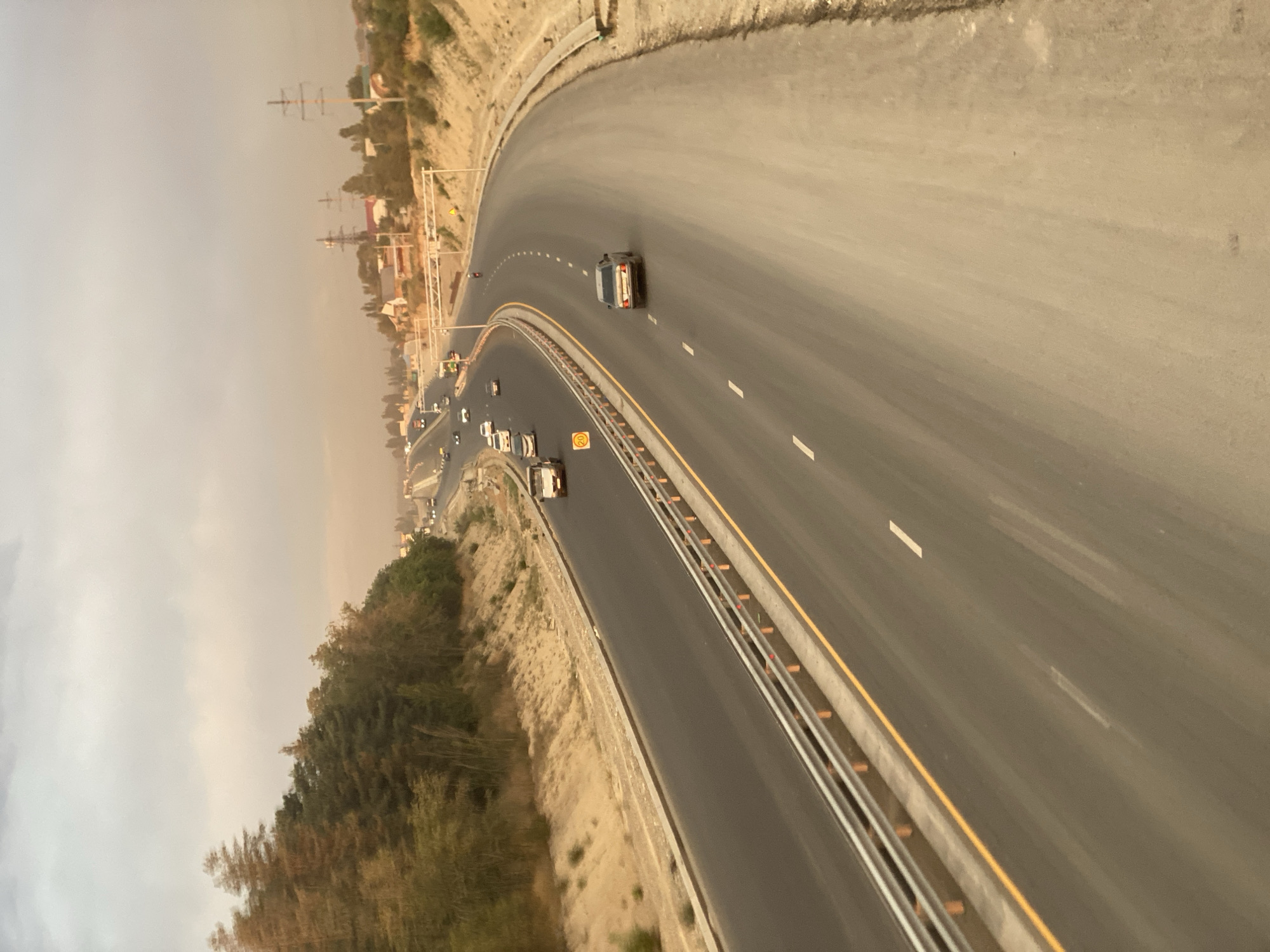 The image captures a view of a multi-lane highway with moderate traffic, likely on the outskirts of Bishkek, Kyrgyzstan. The highway curves to the right and is flanked by dry, sandy embankments and sparse vegetation, including trees on the left side. Power lines and some residential structures are visible in the distance on the right. Several cars and a truck are driving on the highway. The sky is overcast with a hazy, dusty appearance, suggesting potentially poor air quality or a specific atmospheric condition. The lighting indicates it is daytime, possibly late afternoon. The road surface appears to be asphalt, with clear lane markings.