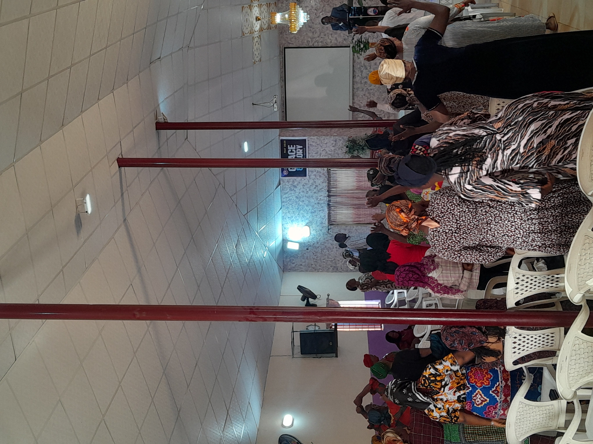 The image shows a group of people gathered in a room, appearing to be praying or in a worship setting. They are sitting on white plastic chairs. The location is in Jos, Nigeria. Several people are facing forward with their hands raised. There is a man standing in the front. A white screen hangs on the wall. Red metal support poles are distributed throughout the room. A sign with the words "Grace Glory" is visible.