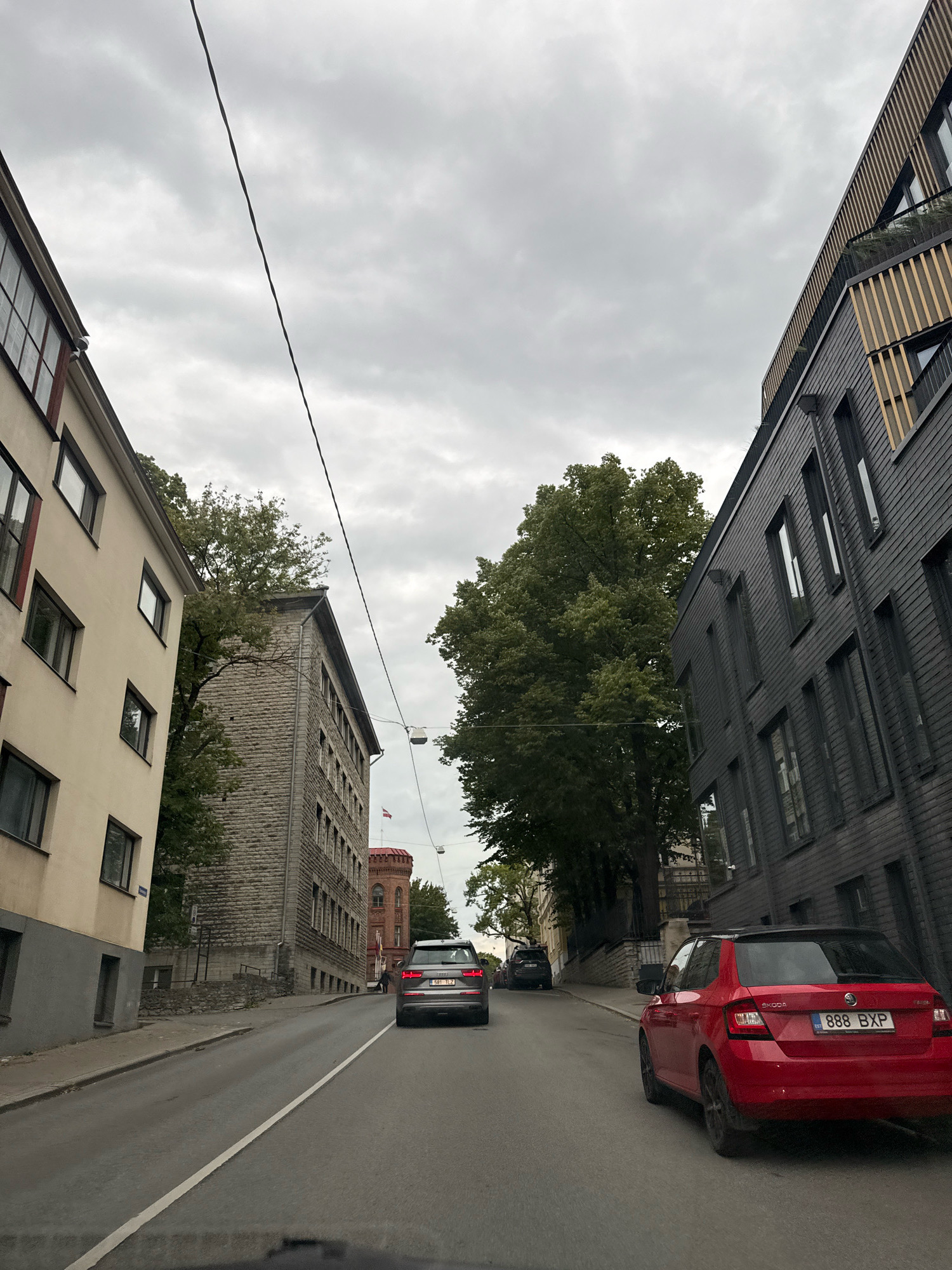 The image shows a street scene in Tallinn, Estonia, under an overcast sky. In the foreground, a red Skoda Fabia hatchback with license plate 888 BXP is parked on the right side of the road, facing away from the viewer. To the left, an Audi SUV is driving away from the viewer. The road is paved and has a white line marking the lane.
On the right side of the street, a dark-colored building with horizontal siding and multiple windows is visible, its upper floors featuring wooden accents and balconies. On the left side of the street, a light-colored building with several windows and a darker grey base is present. Further down the street, a light grey, stone-clad building with multiple rows of windows stands prominently. In the background, a red tower with a flag is visible, and lush green trees line the street, with their branches extending over the buildings. A power line stretches across the sky. The scene appears to be a residential or commercial area within the city.