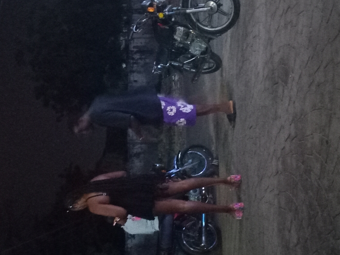 Two people stand on a paved surface near motorcycles. One person wears a dark shirt and purple shorts with a floral pattern. The other individual wears a dark dress and pink sandals. The setting appears to be outdoors at night or in low light. A wall or structure is visible behind the people and motorcycles.