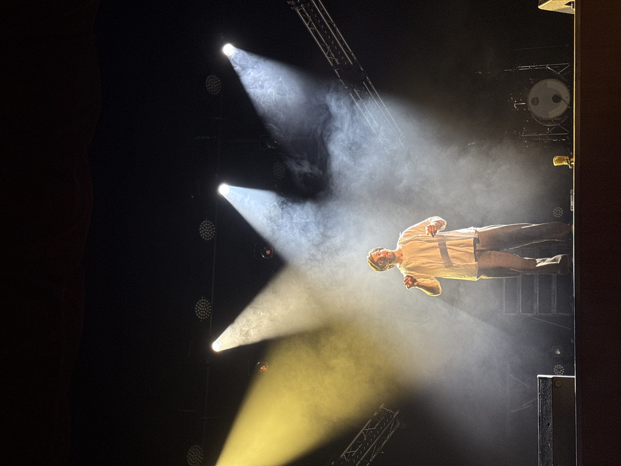 A man in a light-colored t-shirt and dark pants is on a stage, illuminated by spotlights and smoke. The man appears to be performing, possibly singing or speaking, with his body angled and his arm raised. The stage setting includes stage lighting, rigging, and a drum kit in the background. The overall mood is dramatic and energetic, typical of a live performance. The image was taken in Perm, Russia, during a performance or concert.