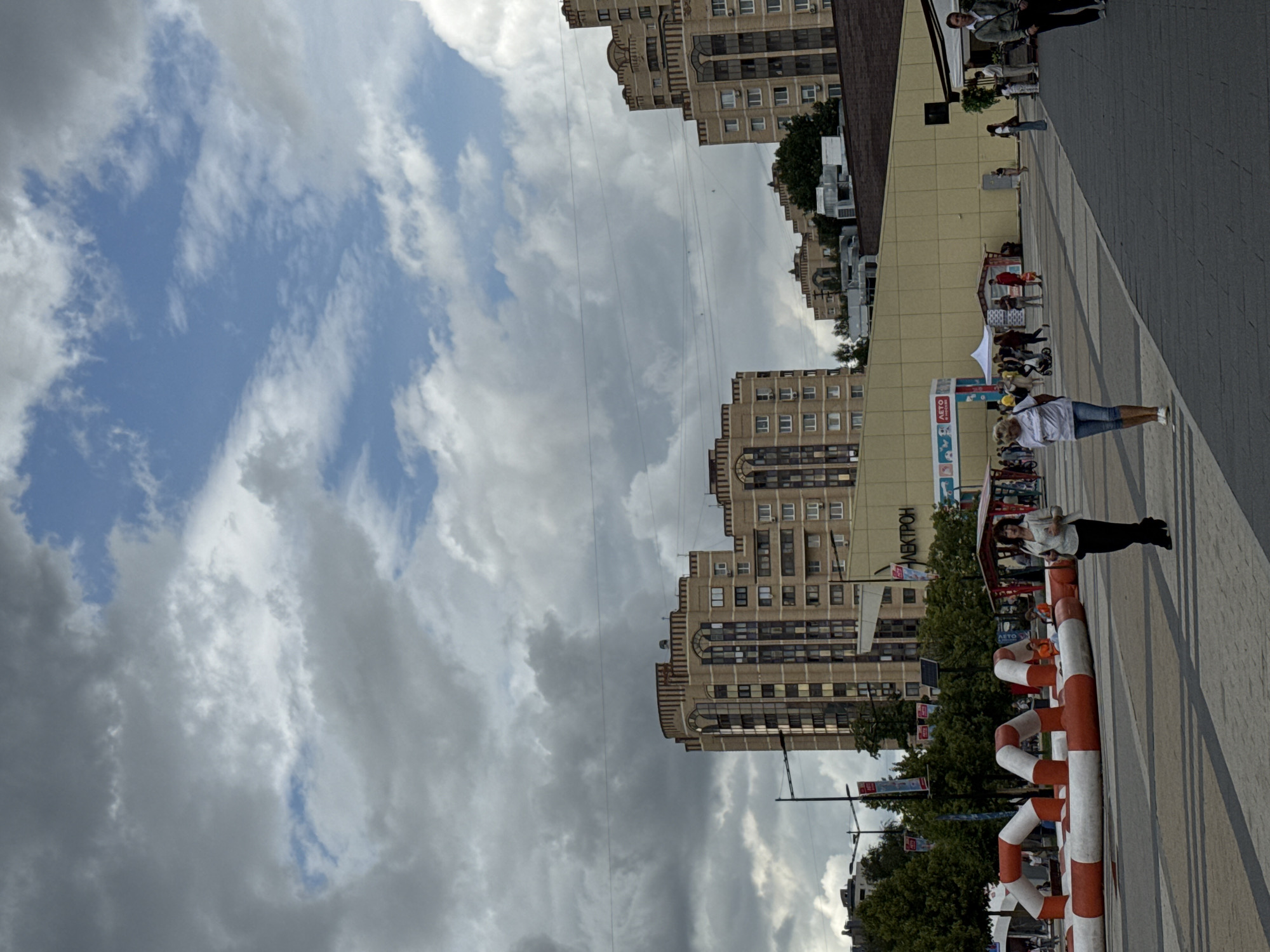This is a medium-angle, eye-level shot of an urban outdoor public space in Zelenograd, Russia, under a cloudy sky with patches of blue. In the foreground, a paved area is visible, with an inflatable obstacle course in the mid-left, featuring red and white tubular structures. Two women are walking across the paved area. One woman in the foreground right wears a white top and black pants. Behind her, another woman in a white t-shirt and blue jeans walks away from the camera.
In the mid-ground, a long, yellow-painted building runs horizontally, with a sign that reads "ЭЛЕКТРОН" (ELEKTRON) in large letters on its facade. Several market stalls and tents are set up in front of this building, with people browsing and interacting. A few trees are visible behind the market area.
The background is dominated by tall residential buildings. The closest building on the left is a light-brown, multi-story apartment complex with a distinctive architectural style, featuring arched windows and decorative vertical elements. Further to the right, more apartment buildings of similar design are visible, with some of them appearing slightly older and darker in color. Overhead, power lines stretch across the sky. The overall atmosphere suggests a lively public gathering or market event occurring on a day with mixed weather.
