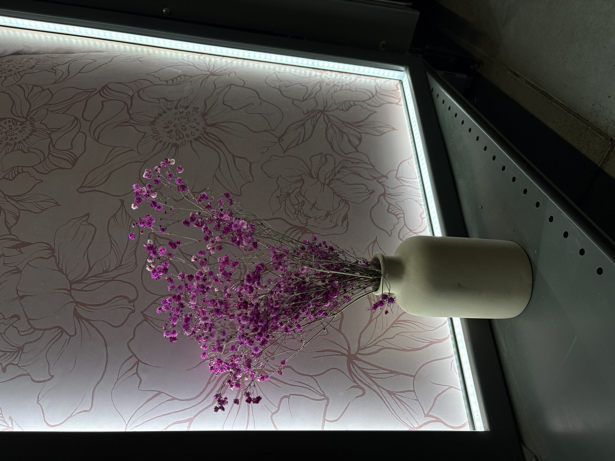The image displays a still life composition, rotated 90 degrees clockwise. In the foreground, a light-colored, possibly ceramic or concrete, cylindrical vase with a narrow neck holds a generous bouquet of dried purple flowers, resembling baby's breath. The small, delicate blooms create a dense, textured mass.
The vase and flowers are positioned against a rectangular, backlit panel that serves as the background. This panel features an elegant floral pattern, consisting of outlines of various large flowers and leaves, rendered in a light pink or reddish-brown hue against a lighter, possibly off-white or very light pink, surface. Bright LED strip lights are visible along the top and left edges of this panel, providing strong illumination to the background.
The entire setup appears to be framed by a dark, possibly metallic, structure. To the right of the backlit panel, there is a darker gray or black surface with a series of regularly spaced circular perforations or indentations, suggesting a functional or decorative element of a larger structure. Beyond this, the background recedes into a dark, undefined area.
The scene is an indoor, static display, likely decorative. There are no people present, and no activity is taking place. The artificial lighting makes the time of day indeterminate. No specific location cues are visible within the image itself to confirm the provided context of Yekaterinburg, Russia. There is no visible text. The overall impression is one of calm, contemporary aesthetics.