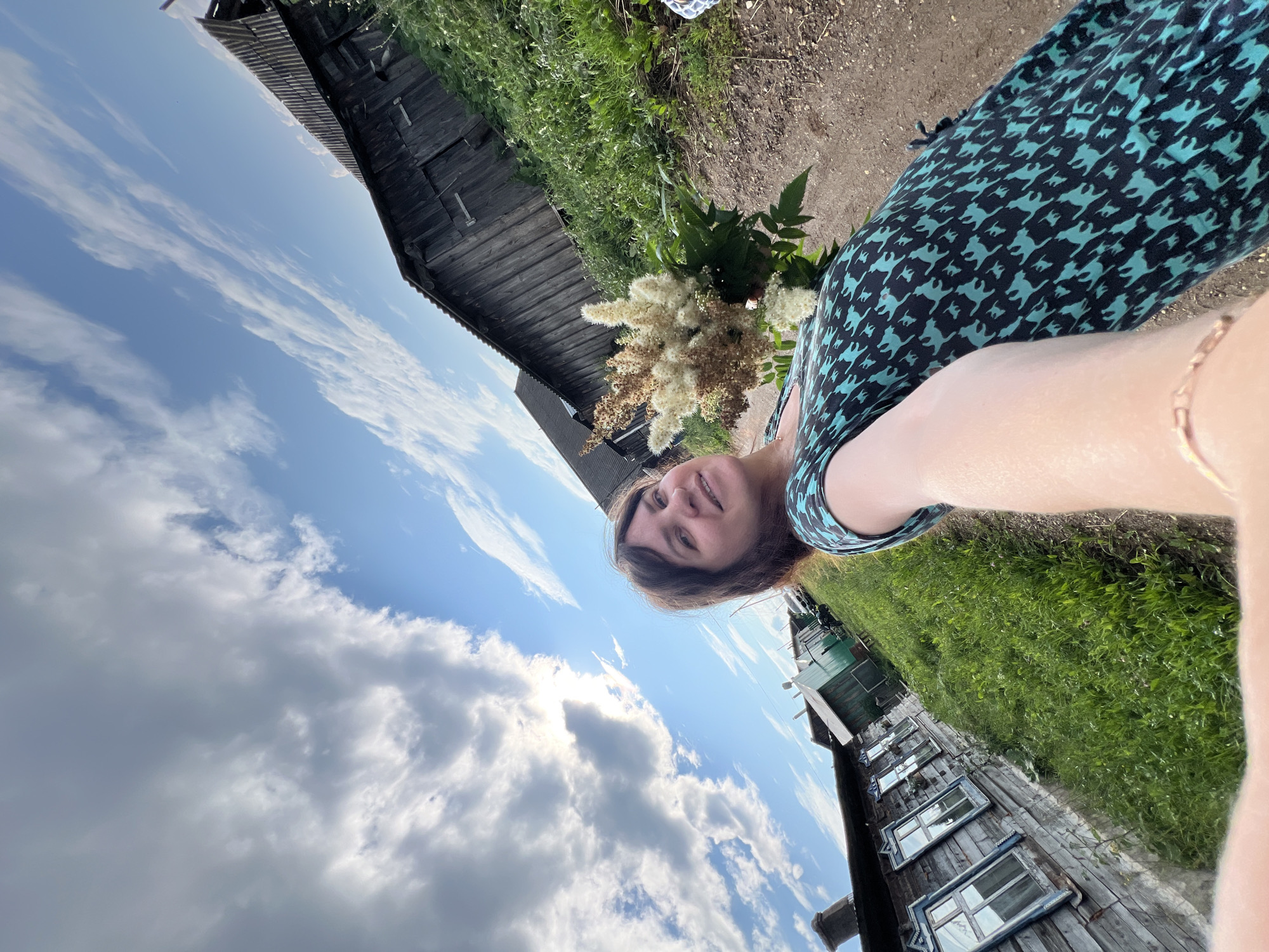 The image depicts a woman taking a selfie in a rural setting, likely in the city of Panteleevka, Russia. The woman is holding a bouquet of white and brown flowers and is wearing a teal dress with a black pattern. She is positioned in the foreground, with a dirt path and lush green grass extending behind her.
In the background, several wooden buildings are visible, suggesting a village or rural settlement. Some of these structures appear weathered and rustic, with visible wood grain and metal roofing. The architecture suggests a traditional Russian style. A clear blue sky with scattered white clouds forms the backdrop, with the sun peeking through some of the clouds, creating a bright and airy atmosphere. The overall impression is one of a serene, outdoor scene in a natural, possibly agricultural, environment.