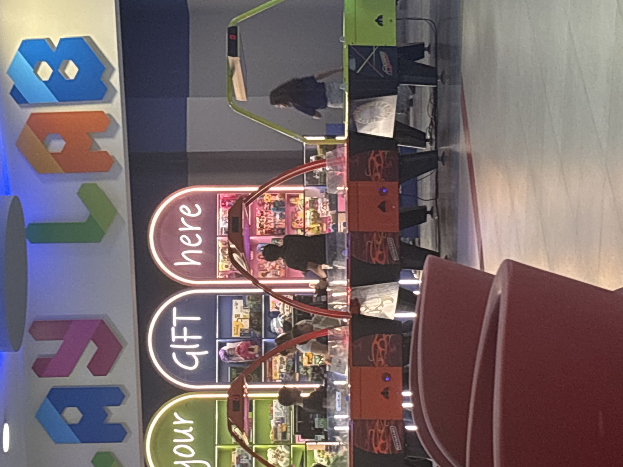 The image, rotated 90 degrees counter-clockwise from its correct orientation, depicts an active indoor arcade or entertainment area, likely located in Yerevan, Armenia. The scene is brightly lit by artificial lights.
On the left side of the frame, a prominent, colorful three-dimensional sign spells out "Toys" using distinct geometric shapes: a blue 't' (or similar block), an orange 'o' (or block with a circular hole), a green 'y' (or block with a V-shape), and a pink 's' (or block with an s-curve). Another green letter is partially visible below the pink 's'.
In the center, a row of arcade machines with illuminated panels displays the words "your", "GIFT", and "here" arranged vertically. These appear to be prize redemption games, possibly claw machines, as various small toys and items are visible inside. One person, wearing a black top, is actively playing one of these machines. The side panel of an orange machine clearly shows the text "NAMCO", indicating the game's manufacturer.
Further back and to the right, another arcade game with a bright green frame is in use. This game resembles an air hockey table, and a second individual, with dark hair and dressed in a dark top, is engaged in playing it. A third person is partially visible behind the prize machines.
On the far right, a sequence of red, curved chairs suggests a waiting or seating area. The floor is light-colored and smooth. The overall impression is one of a lively and engaging recreational space.