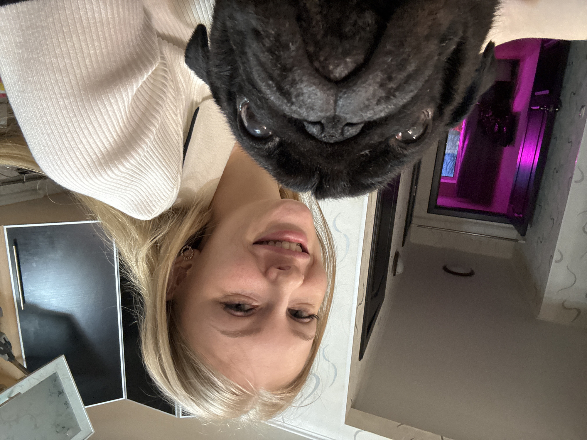 A selfie taken in Perm, Russia shows a blonde woman with fair skin smiling slightly, positioned to the left of a black pug whose face fills the top right portion of the frame. The woman wears a cream-colored ribbed sweater and small, round earrings. The pug's dark, wrinkled face is prominent, with its eyes visible. The background includes a door frame with a purple glow coming from the room behind it and a wall with a subtle pattern. The lower left corner of the frame shows a black surface and a metallic stand.