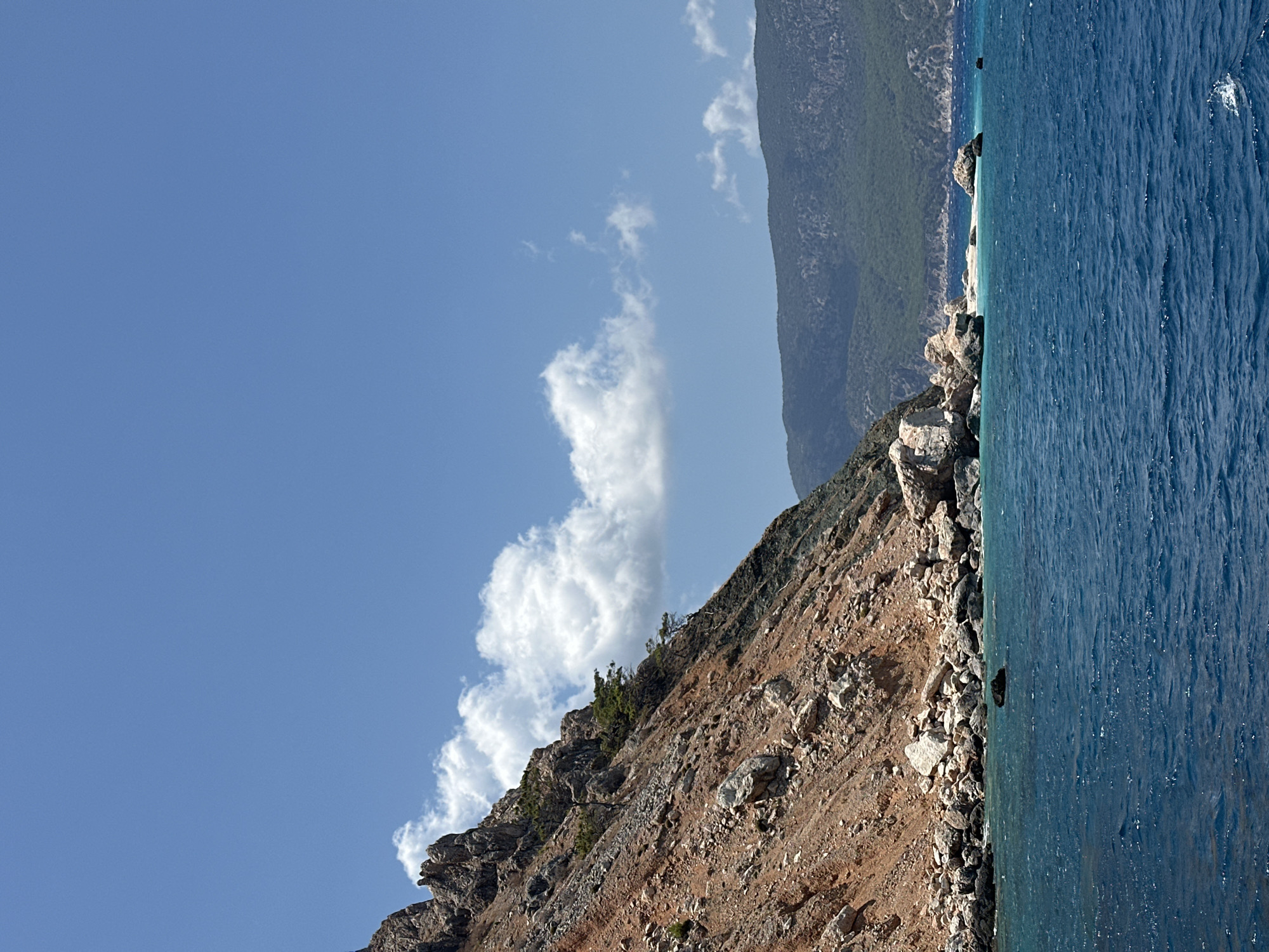 A rocky, reddish-brown mountainside slopes down to a turquoise sea under a bright blue sky with scattered white clouds. The mountain is sparsely populated with trees, some of which are clinging to the steep incline. Rocks of various sizes are strewn across the mountainside and at the base where it meets the water. In the distance, a range of green, forested mountains is visible. The water is choppy, indicating a gentle breeze. There are no people or man-made structures visible in the image. The scene depicts a natural, coastal landscape.