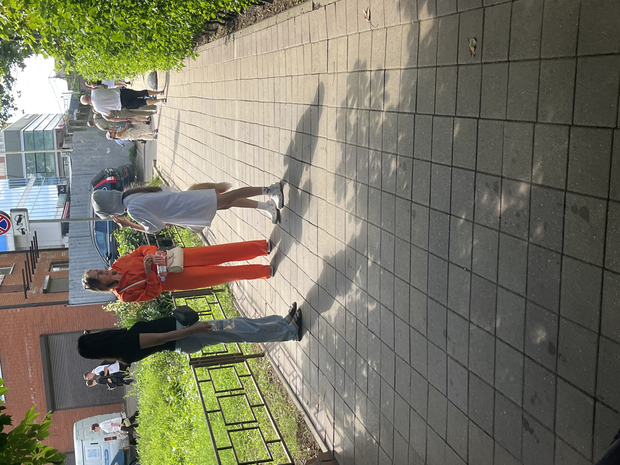 The image shows a sunny day on a paved sidewalk in Krasnodar, Russia. The sidewalk is made of grey square tiles. On the left side of the image, a woman in a black shirt and light blue jeans is standing, holding a plastic bottle. Next to her, a woman in an orange jumpsuit is speaking and holding a plastic bottle and a small purse. Further down the sidewalk, a young girl in a white dress and a grey hat is facing away from the camera. She is holding a fluffy object. In the background, several other people are walking, and there is a black car parked behind a metal fence. A green bush line the right side of the sidewalk, and a brick building is visible behind the people on the left.