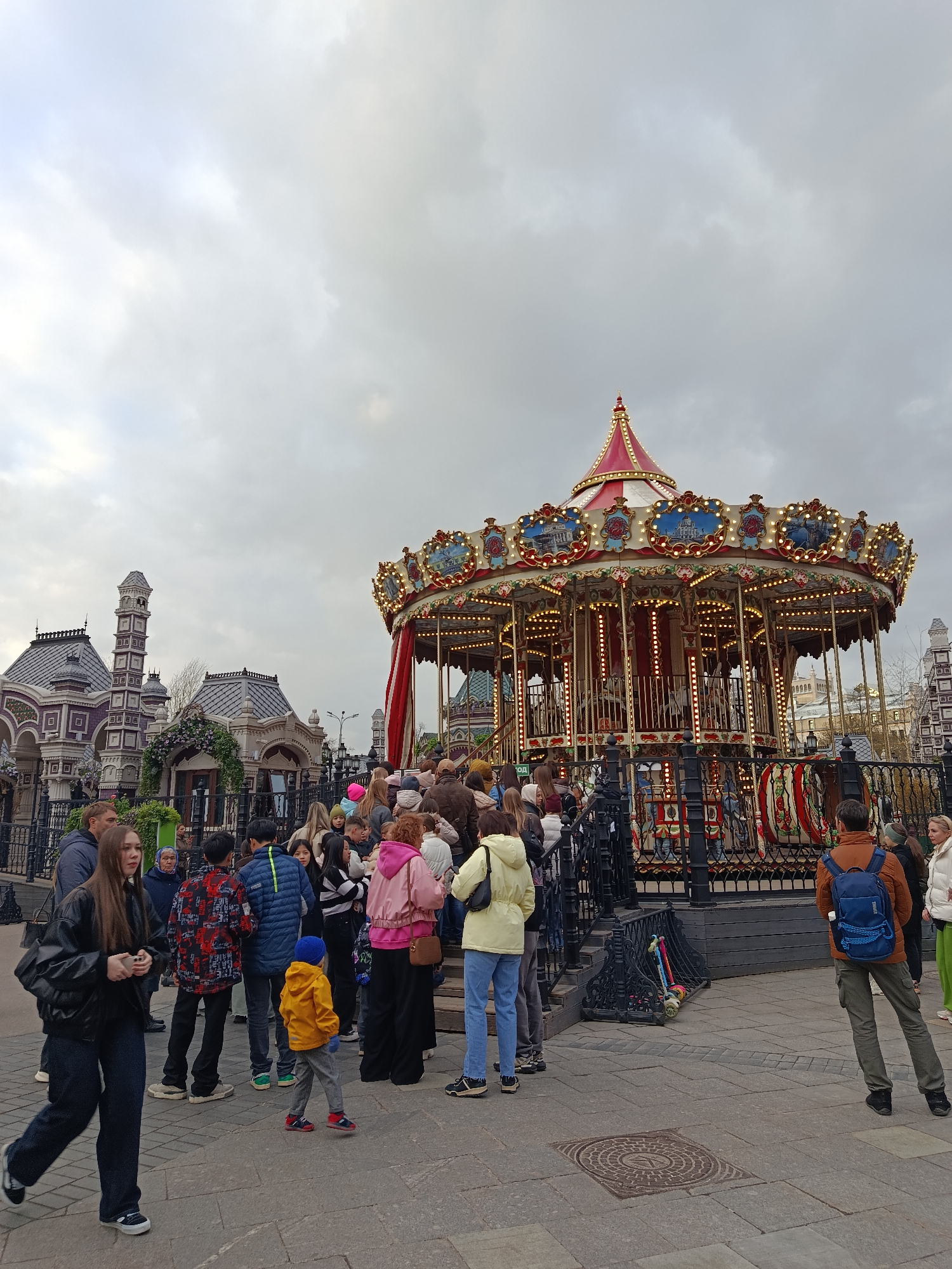 The image captures a bustling outdoor scene featuring a grand, traditional carousel as its focal point. The carousel is richly decorated with red and white striped awnings, gold accents, and numerous small, glowing lights, suggesting it's either evening or a dark, overcast day. Ornate panels depicting scenes are visible along its circumference, and a black wrought-iron fence surrounds its base, behind which many people are gathered, seemingly queuing or watching.
In the background, there are distinctive buildings with intricate, multi-tiered architecture, characterized by dark roofs, decorative facades, and tall, slender towers, hinting at a historical or unique setting. One building on the left has greenery draped over an archway.
A diverse crowd of people occupies the foreground and midground. There's a mix of adults, teenagers, and children, dressed in various casual attire including jackets, hoodies, and jeans, suggesting cool weather. Many individuals are standing near the carousel, while others are walking through the paved plaza. One person on the far right wears an orange jacket and a blue backpack. A child in a bright yellow jacket walks in the lower-middle frame. Scooters or toys are seen leaning against the carousel fence.
The sky above is heavily overcast with light grey clouds. The ground is paved with large, grey flagstones, and a manhole cover is visible in the lower right. A partial sign near the carousel reads "ОД", likely part of "Вход" (Entrance) in Russian. The overall atmosphere is that of a public gathering point, possibly a market, park, or fairground.