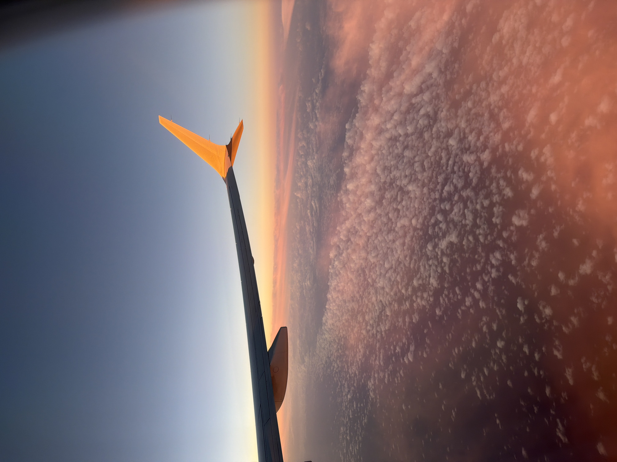 The image shows an airplane wing in flight. The wing is on the left side of the image, and it is angled upwards. The sky is visible in the background, and it is a gradient of colors from blue to orange. There are clouds in the distance. The location is unknown.