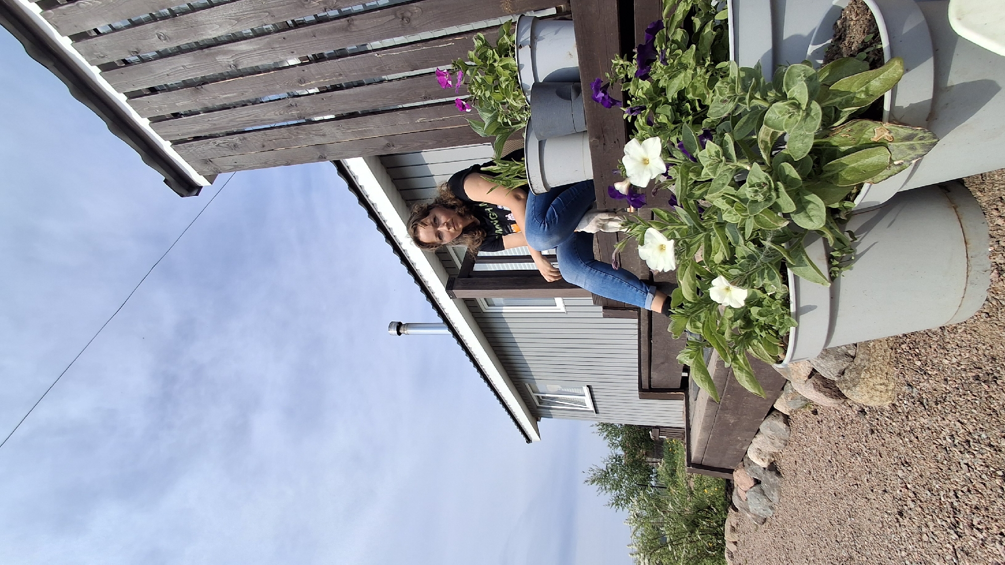 The image captures a woman tending to plants in a multi-tiered planter. The planter is located on a deck next to a house. The house is in Kempelevo, Russia, as suggested by the metadata. The woman is wearing jeans and a dark t-shirt and is bending over, her hands near the plants. The plants feature green foliage and both white and purple flowers. The background includes a gray house with a chimney and a partially visible sky with clouds. The ground around the deck is covered in gravel.