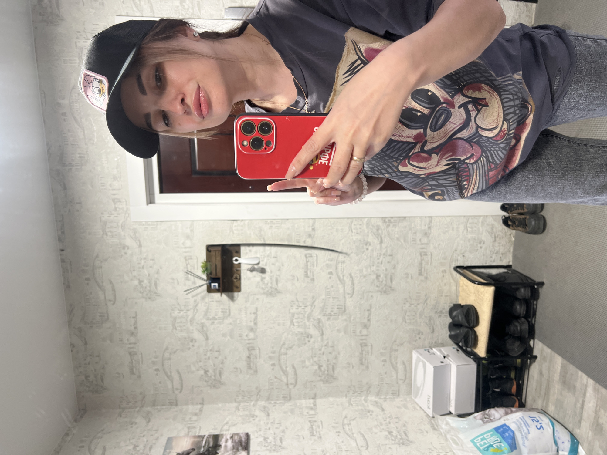 A mirrored view reveals a woman taking a selfie in what appears to be a residential interior. She is wearing a dark t-shirt with a Mickey Mouse-style graphic, dark jeans, and a baseball cap. She holds a red smartphone with a triple-lens camera, possibly an iPhone, in her hand. The mirror reflects a room with patterned wallpaper, a dark door frame, and a white door. In the background, a shoe rack stacked with shoes is visible near the wall. There are also two white boxes near the shoe rack. A dark-colored wall fixture with decorative items is mounted on the wall. A white baseboard runs along the bottom of the wall. A partial view of a framed print or photograph can be seen on the floor. The location is Unknown, Unknown.