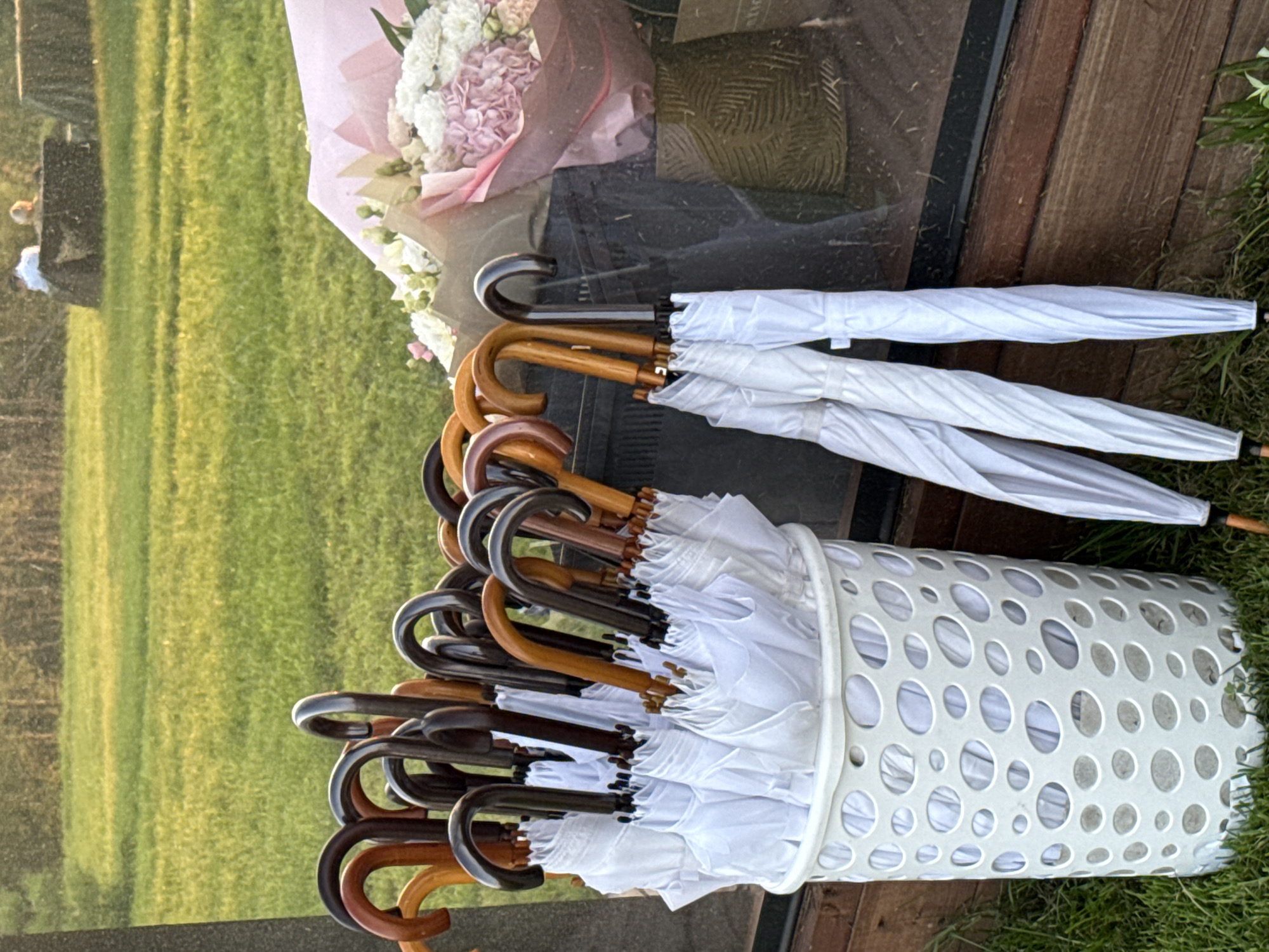 The image depicts an outdoor scene with a focus on a collection of umbrellas and a floral arrangement. In the foreground, a white, perforated plastic basket overflows with numerous white umbrellas. The umbrellas are mostly closed, with their wooden handles, in various shades of brown and black, visible at the top. To the right of the basket, three more white umbrellas are leaning against a dark surface, their handles also visible. Behind the umbrellas, a large bouquet of flowers, predominantly pink and white, is partially obscured by a dark material. The bouquet appears to be wrapped in pink paper. The background features a green grassy field, with trees visible in the distance. The lighting suggests it is late afternoon or early evening, with a warm, golden hue cast over the scene. There are no people clearly visible in the image. The location is not specified to be in the city of Светлое, Russia.