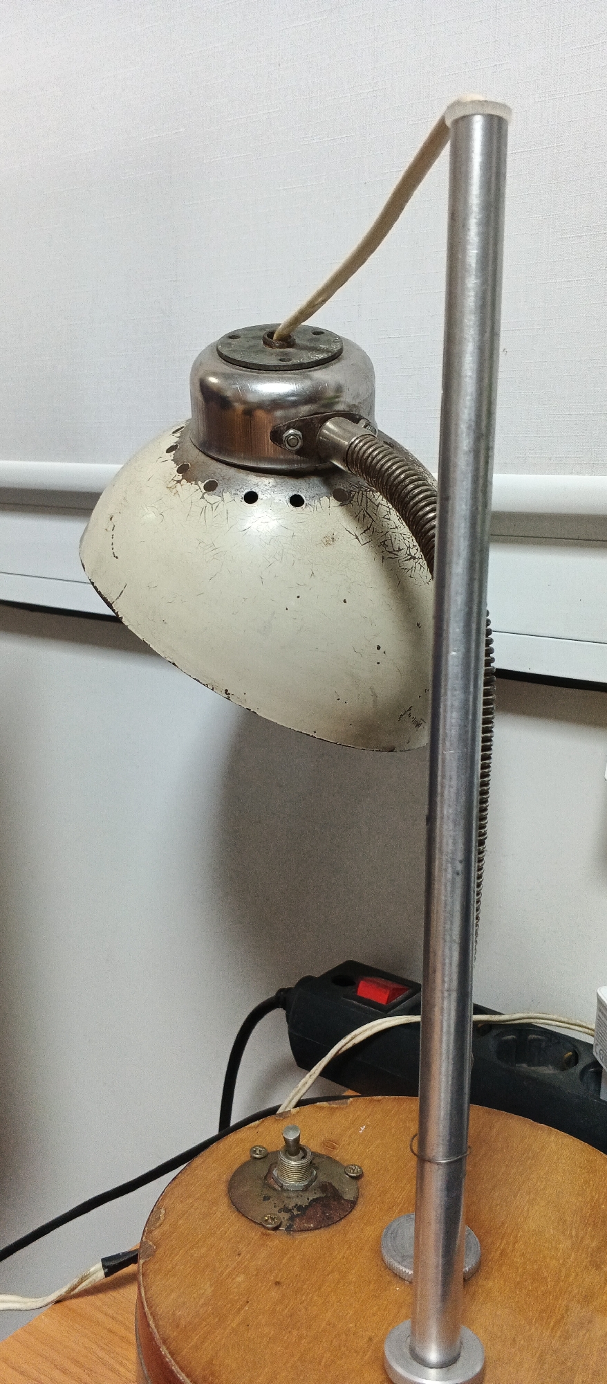 A close-up shot focuses on the base and upper portion of an old-fashioned, metal desk lamp with a cream-colored, conical shade. The lamp is situated on a round, wooden base. A silver, cylindrical pole rises from the base, supporting the lamp head. The lamp shade shows signs of wear, with chipped paint revealing a darker layer underneath. A coiled metal arm connects the shade to the pole. A light-colored cord emerges from the top of the lamp's metal housing and loops over the silver pole before disappearing out of frame.
In the background, a white wall with a subtle texture is visible, along with a horizontal blind. A black power strip with a red switch is located near the base of the lamp, with power cords running from it. The overall environment appears to be indoors, possibly a workshop or study, but no specific location in the city of Smila, Ukraine, can be identified from the image. There are no people or animals present in the scene. The lighting suggests an indoor setting, possibly illuminated by ambient light or the lamp itself (though it is not illuminated in the photo).