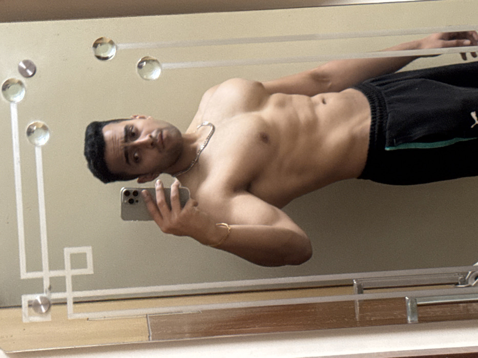 A shirtless man with dark hair and a silver necklace takes a selfie in a mirror. He wears black shorts with a green stripe down the side. His bare torso is visible, showcasing his muscular physique. The mirror has a decorative frame with circular accents and geometric patterns. The background is a plain, light-colored wall. The bottom of the mirror rests on a wooden surface.