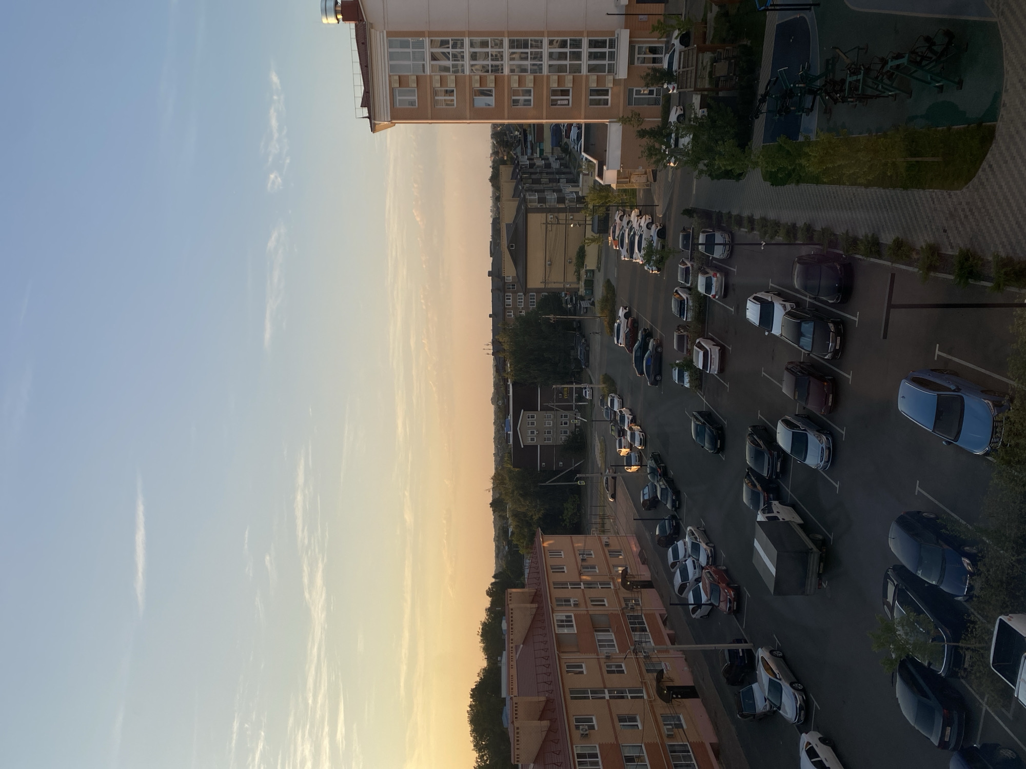 This is an aerial, full-frame photograph taken at dusk, looking down on a residential area with apartment buildings and a parking lot. The sky is a gradient of pale blue at the top transitioning to a warm orange and yellow near the horizon, with scattered wispy clouds. In the foreground and to the right is a modern, multi-story apartment building with light-colored walls and numerous windows. To the left is another apartment building with a similar color scheme and red-tiled roofs.
The central focus of the image is a paved parking lot filled with numerous cars of various makes and models, parked in designated spots. The cars are mostly dark in color, including black, dark blue, and silver. There is a white truck with a grey cargo box parked in the middle of the lot. A few people are visible walking in the distance, near the apartment buildings.
Beyond the immediate apartment complexes, there are more buildings and trees, suggesting a suburban or urban residential setting. The overall impression is a quiet evening in a developed neighborhood, likely in Vishnyaki, Russia, given the architectural style and the presence of diverse car models common in the region. The lighting suggests the sun has recently set, casting long shadows and bathing the scene in a soft, warm glow. To the far right of the image, a small outdoor exercise area with equipment is visible.