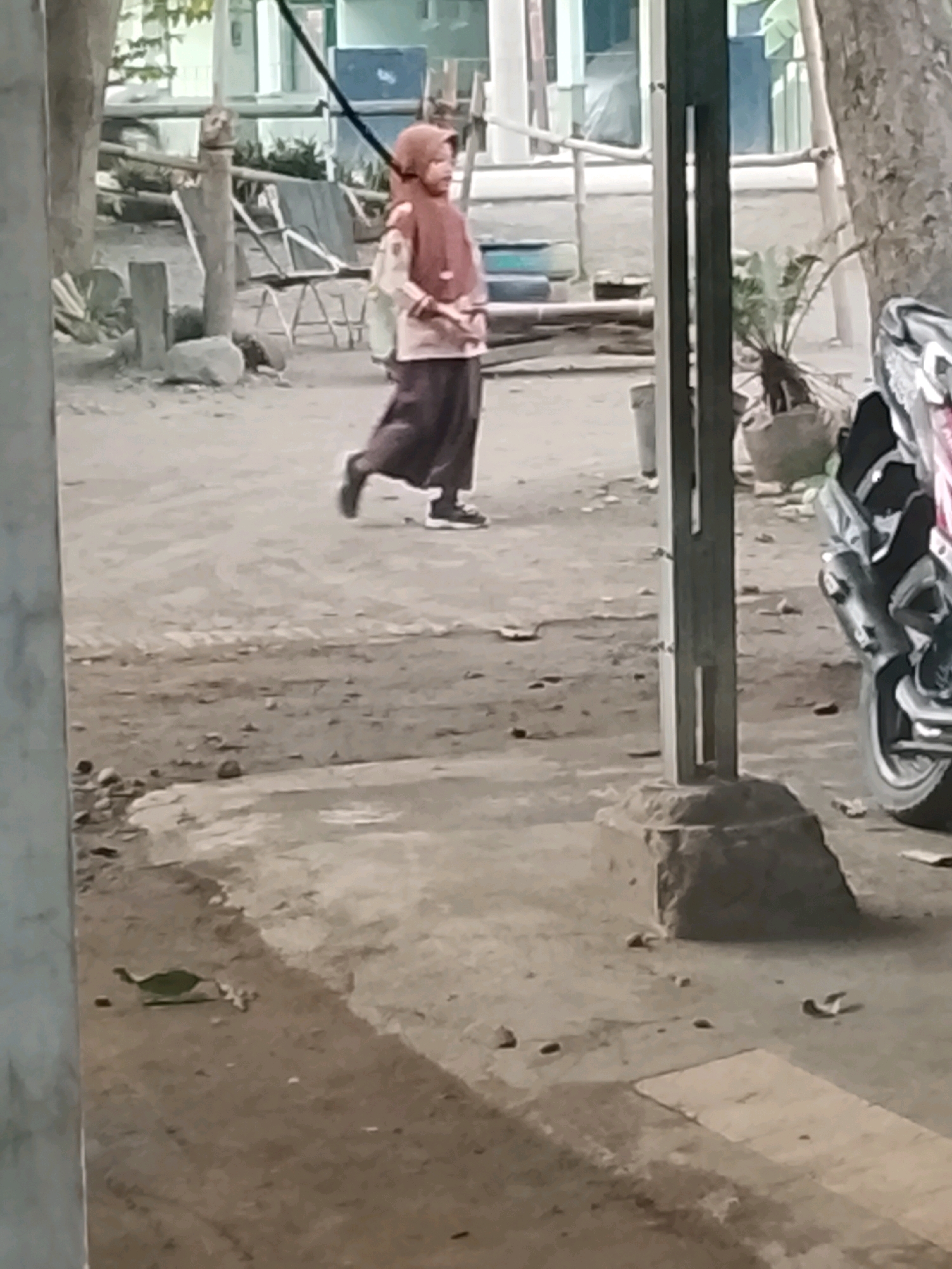 A child wearing a headscarf and long dark skirt walks to the right across a dusty outdoor area. The child's face is not visible. A motorcycle is parked on the right side of the image. The background shows trees and buildings. The scene is likely in Ujungrusi, Indonesia, given the context provided.