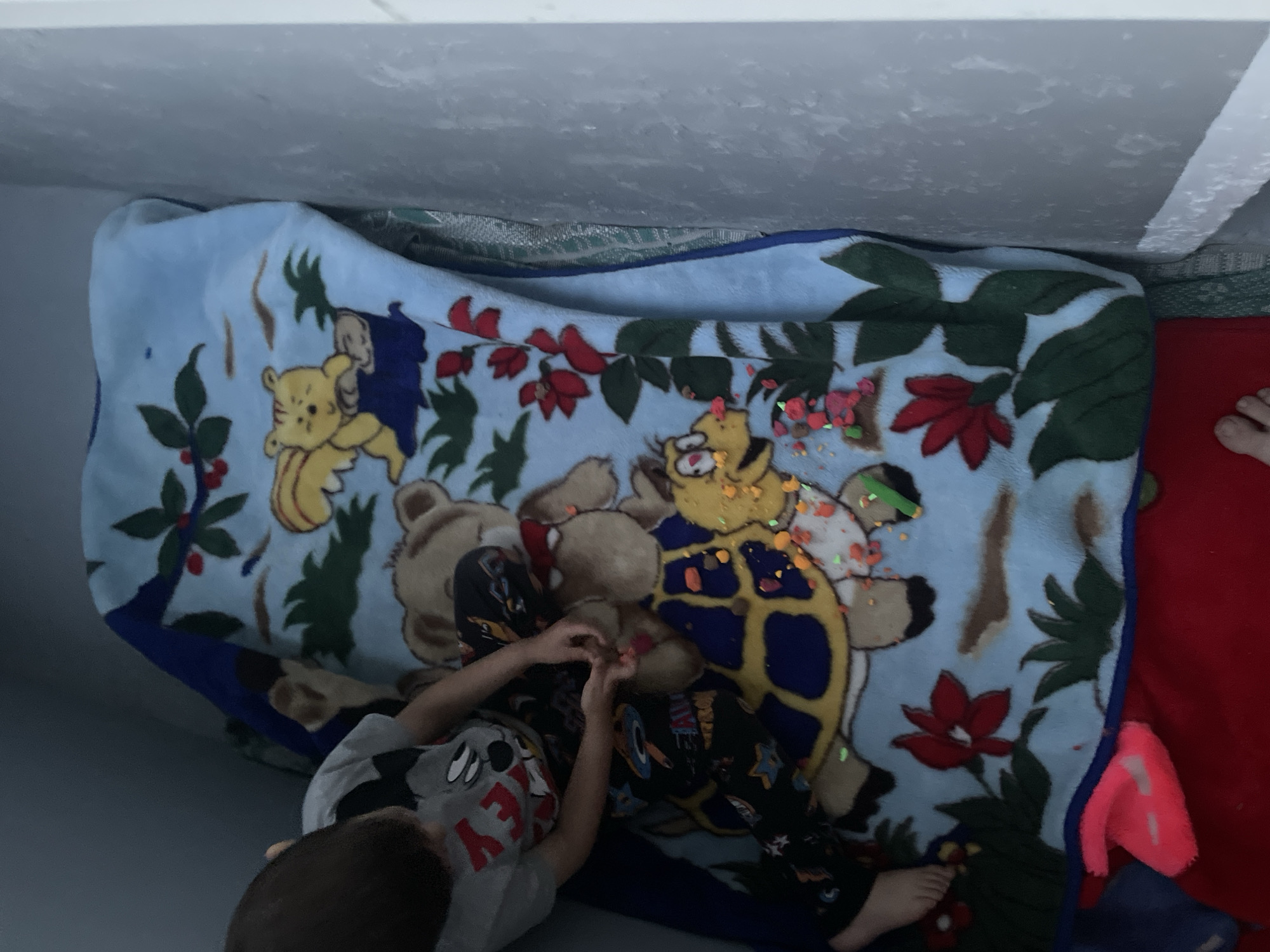 The image shows a young child sitting on a bed covered with a light blue blanket featuring cartoon characters of a bear and a turtle. The child is wearing a grey t-shirt with a Mickey Mouse graphic and patterned pajama pants. The child's hands are interacting with small colorful objects scattered on the blanket, specifically on the turtle's shell. The objects appear to be small candies or beads. The bed is positioned in a corner, with a grey wall on the right side of the frame. A pink object is visible at the bottom left of the image, partially obscured by the blanket. The lighting suggests an indoor setting, possibly a bedroom.