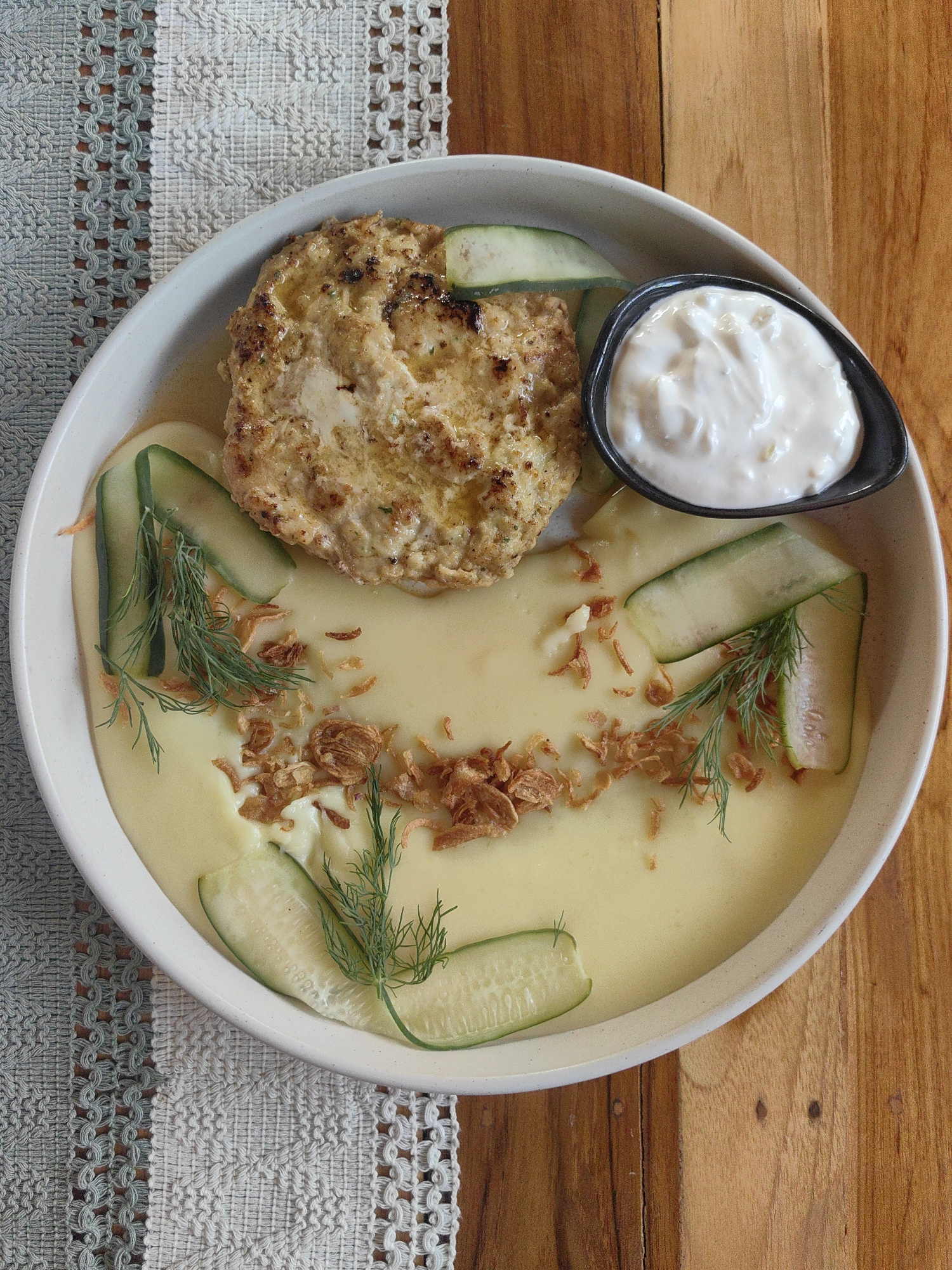A close-up, overhead shot shows a white bowl filled with mashed potatoes, topped with a golden-brown patty, cucumber slices, dill sprigs, and fried shallots. A small black dish contains a dollop of white sauce, likely sour cream or yogurt, situated to the upper right of the patty. The bowl is placed on a woven placemat with a patterned design, which lies on a rustic wooden table. The lighting suggests daytime, possibly indoors or in a shaded outdoor area. The setting appears to be a restaurant or dining establishment, given the presentation of the food and the presence of a placemat. No people are visible in the image. The food looks ready to be served or eaten. The scene conveys a sense of a prepared meal, likely a main course. The overall impression is one of a well-presented dish in a comfortable, possibly casual dining environment.