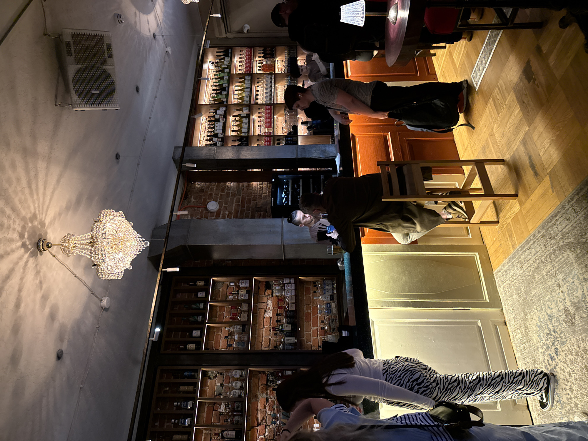 The image depicts the interior of a bar or restaurant, possibly located in Moscow, Russia, given the architectural styles sometimes found there. The setting includes a mix of exposed brickwork and painted walls. There are several people visible in the scene. Some appear to be patrons seated at a bar or tables, possibly engaged in conversation or looking at their phones. A prominent feature is the shelving behind the bar, displaying an extensive collection of liquor bottles. Overhead lighting includes a large, decorative chandelier and other light fixtures above the bar area. There's a speaker mounted on the upper portion of the wall. The floor is a light wood parquet. Some of the people are partially obscured or cut off by the frame of the image. There is an area rug on the floor.