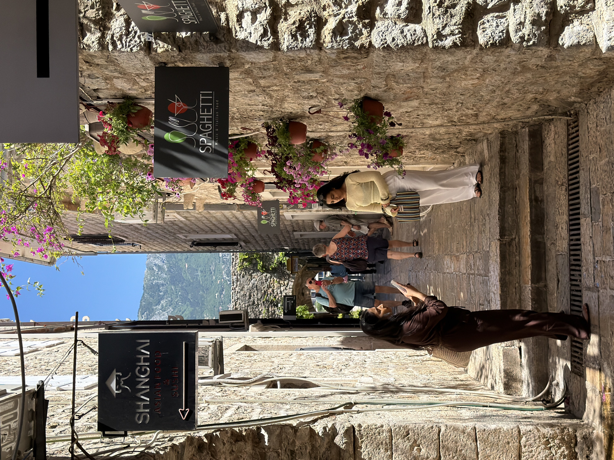 Captured in the city of Budva, Montenegro, is a narrow, cobblestone pedestrian walkway situated between aged stone buildings. People stroll through the walkway. A woman in a brown outfit is on the bottom, right side of the photo, holding a phone. Further up the walkway are a couple, with the man drinking from a can or bottle, and the woman is wearing a patterned dress and carrying a brown bag. A woman in a light yellow shirt and white pants is further up the alley.
Signage indicates "Shanghai, Asian food and Sushi" on the left side of the walkway, and "Spaghetti, music and Italian food" on the right, with potted plants adding color. The sky and mountain backdrop are visible at the end of the alley.
