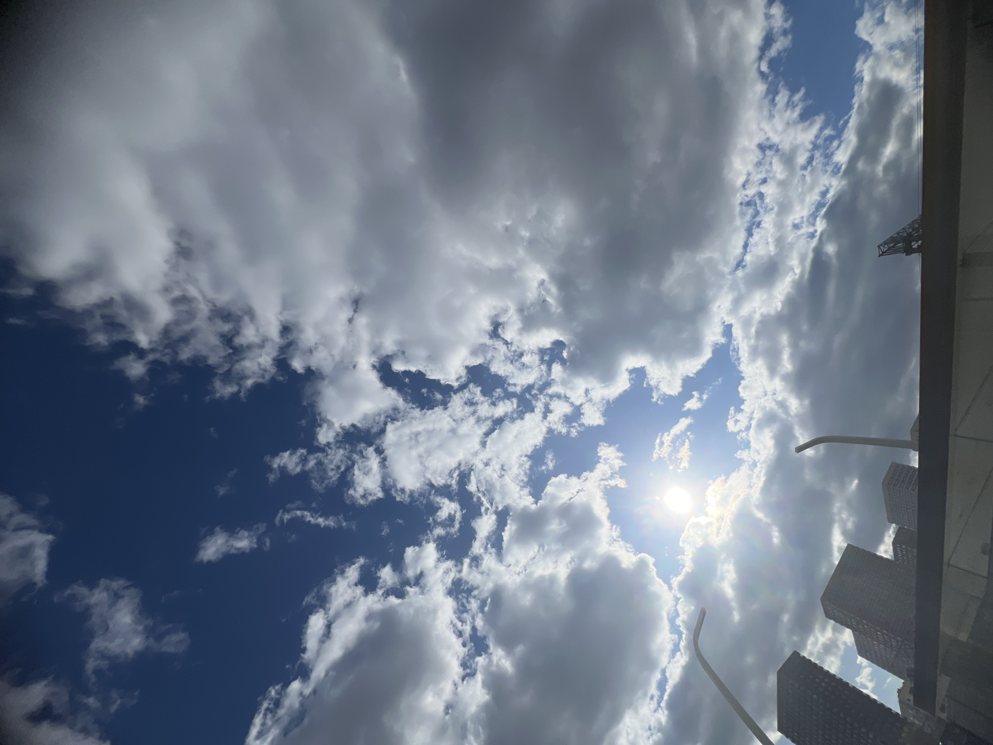 The image is a low-angle shot looking up at a sky with a mix of cumulus and cirrus clouds. Some patches of blue sky are visible. The sun is visible through a gap in the clouds. In the bottom right corner, there is a section of buildings and what appears to be street lights. The photo may not be in Khimki, Russia because there is no evidence in this image to indicate it is from Khimki, Russia.