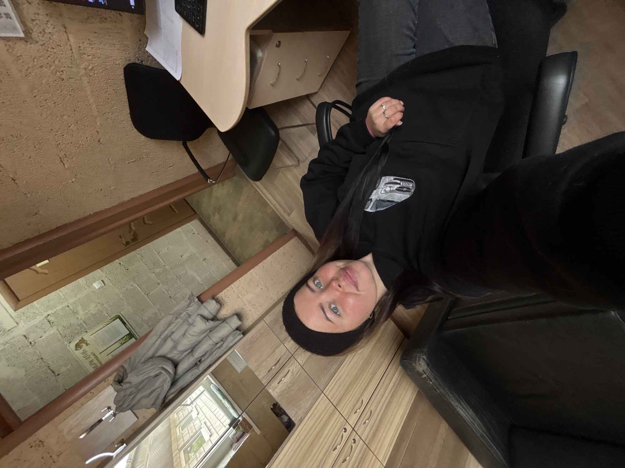 A young woman with long, dark hair and light-colored eyes is seated in a black office chair, looking directly at the camera. She wears a black beanie and a black hooded sweatshirt with a graphic of a car and the word "LIVERY" visible on its front. Rings are worn on her left hand.
The setting is an indoor room, appearing to be a functional workspace or office. To her side is a light-colored wooden desk with multiple drawers, equipped with a keyboard and various papers. In the background, a gray puffer jacket is draped over a wooden railing or bar, and a light fixture is mounted on a wall made of light-colored cinder blocks. On this cinder block wall, a poster or document with Cyrillic text is visible, including the words "РУСЬ 1716" and partially "НИМАЕТ".
A window allows natural light into the room, revealing a view of buildings and some greenery outside, indicating it is daytime. The room features a mix of wall finishes, including the cinder blocks, a textured beige wall, and light wooden paneling below the window. The floor is made of light-toned wood. The overall impression is a casual, somewhat rustic, or utilitarian environment. The woman's expression is calm and neutral. The choice of clothing, including the beanie and puffer jacket, suggests a cooler climate, aligning with the provided location context of Русский Вожой, Russia.