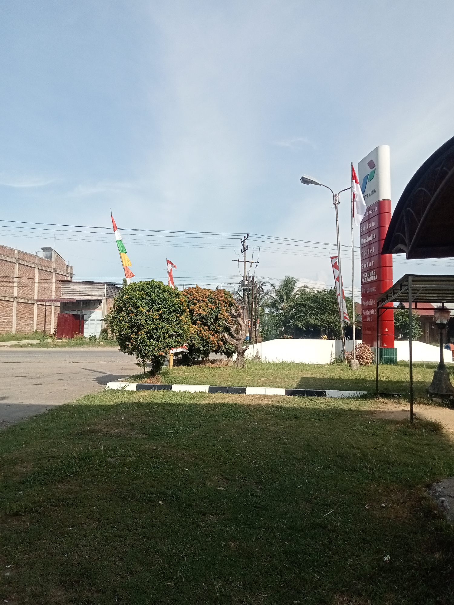 The image captures a view of a Pertamina gas station in Matangpanyang, Indonesia, on a sunny day. In the foreground, a grassy area with a black and white bordered edge is visible. To the right, a covered structure with a lamp post stands. A prominent red and white Pertamina sign is a central element, displaying fuel types and operating hours. Indonesian flags, red and white, are flown from poles near the gas station. Behind the gas station, trees and palm trees form a backdrop. In the distance, a brick building with a corrugated metal roof and a large gray garage door is situated next to a road. Electrical poles and wires are also present in the scene. The sky is clear with a few wispy clouds.