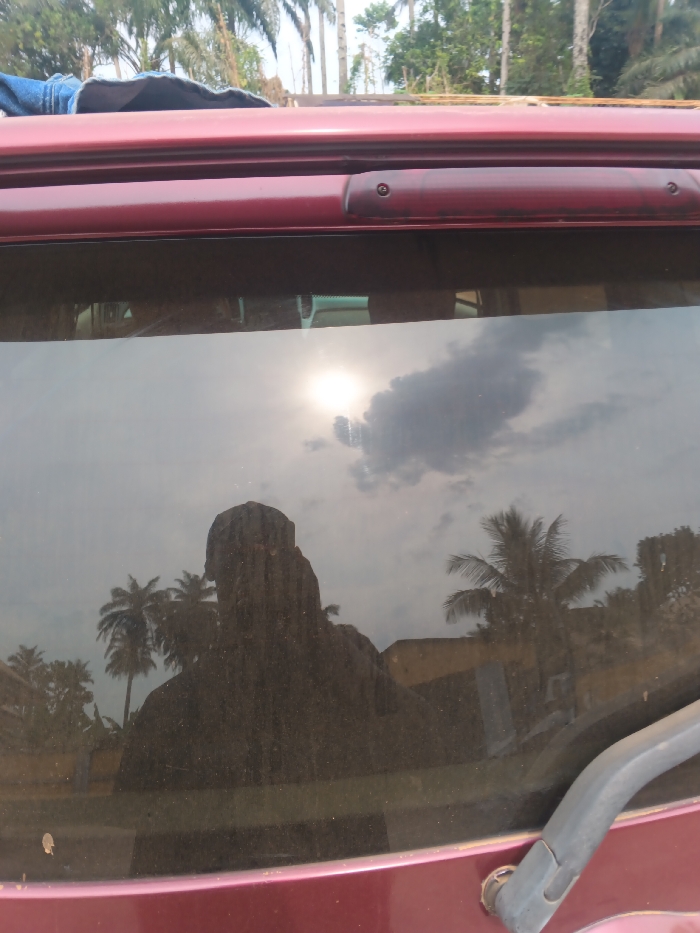 The image depicts an outdoor scene in Itu, Nigeria, viewed through the dusty rear window of a dark red vehicle. The vehicle's dark red body panel forms the bottom and top edges of the frame. A gray rear windshield wiper arm is present in the bottom right corner. Along the top edge of the rear window, a dark red third brake light assembly is visible, fixed to the vehicle's roofline. A folded blue denim-like garment rests partially on the vehicle's roof, visible in the upper-left.
The rear window's surface reflects the immediate surroundings and allows a partial view through it. A prominent, dark silhouette reflection of a person, positioned centrally in the lower half of the window, faces away from the viewer. The reflection also includes a bright sun in the upper-left quadrant of the window, partially obscured by a dark cloud. Other scattered clouds are visible against a lighter sky. Through the window and as reflections, numerous palm trees and other green deciduous foliage are discernible. Reflections of light-colored, possibly single-story buildings are visible in the middle-ground and background. The scene is illuminated by daylight, suggesting a sunny to partly cloudy sky.