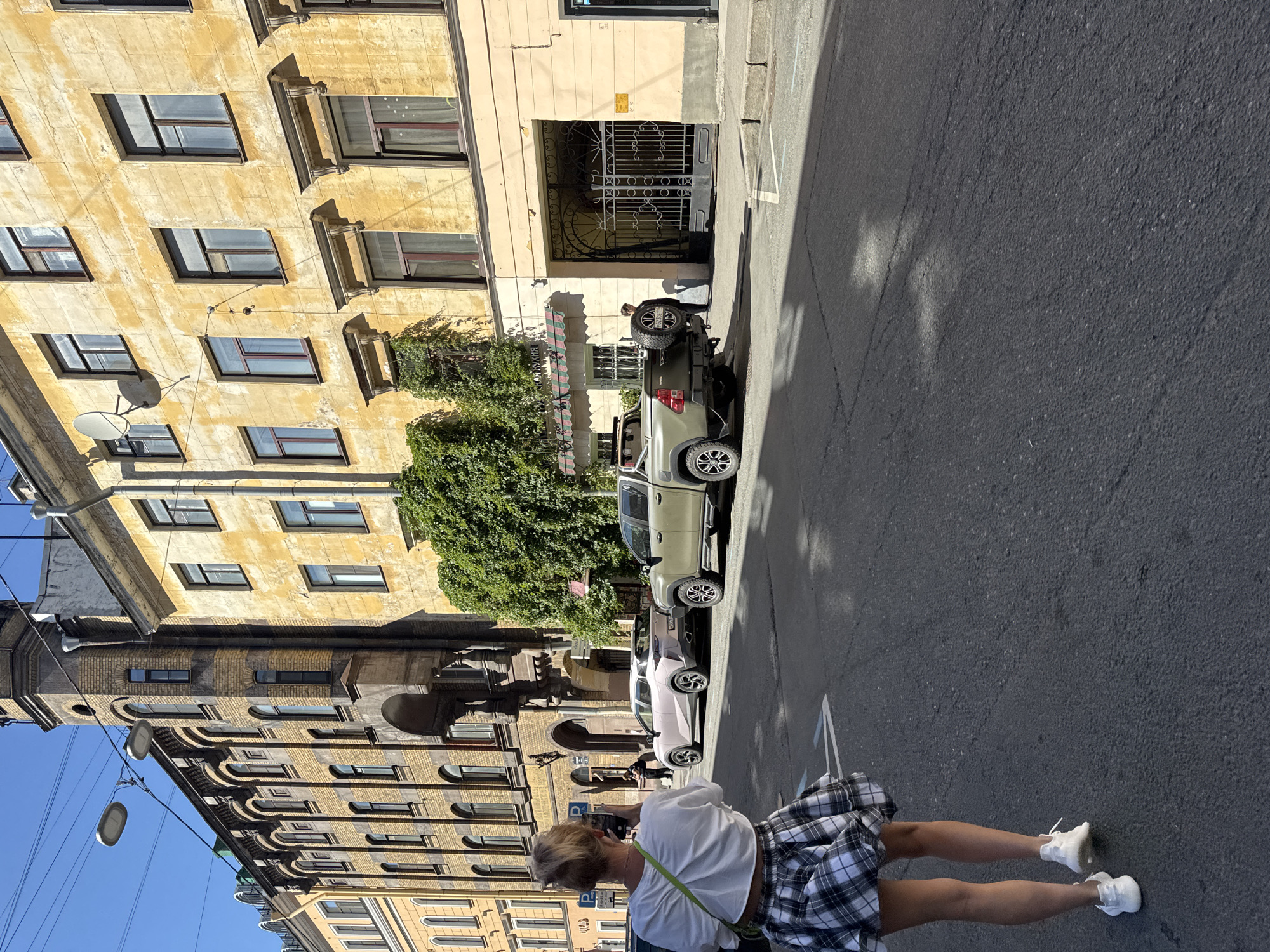 The media file depicts a street scene in Saint Petersburg, Russia, on a sunny day. The focus is on a woman with blonde hair, wearing a white top and a plaid skirt, standing on a paved street. She is captured from behind, with her legs and lower torso visible as she stands with her feet slightly apart. Her right arm is bent, and she appears to be holding a phone, possibly taking a picture.
In the background, several parked cars are lined up along the street. The first car is a light-colored pickup truck with a spare tire mounted on the back. Following it are a few other vehicles, including a white sedan.
The setting is characterized by old, ornate buildings flanking the street. The building on the left features a yellow facade with numerous windows and architectural details. Further down the street, another building with a brick facade and arched windows is visible. Lush green trees and ivy grow on the side of one of the buildings, adding a touch of nature to the urban landscape. An ornate metal gate is prominent in front of one of the buildings.
The sunlight suggests it is daytime, likely mid-morning or afternoon, casting shadows on the street. The overall impression is one of a casual urban stroll in a historic European city. No discernible text is visible in the image.