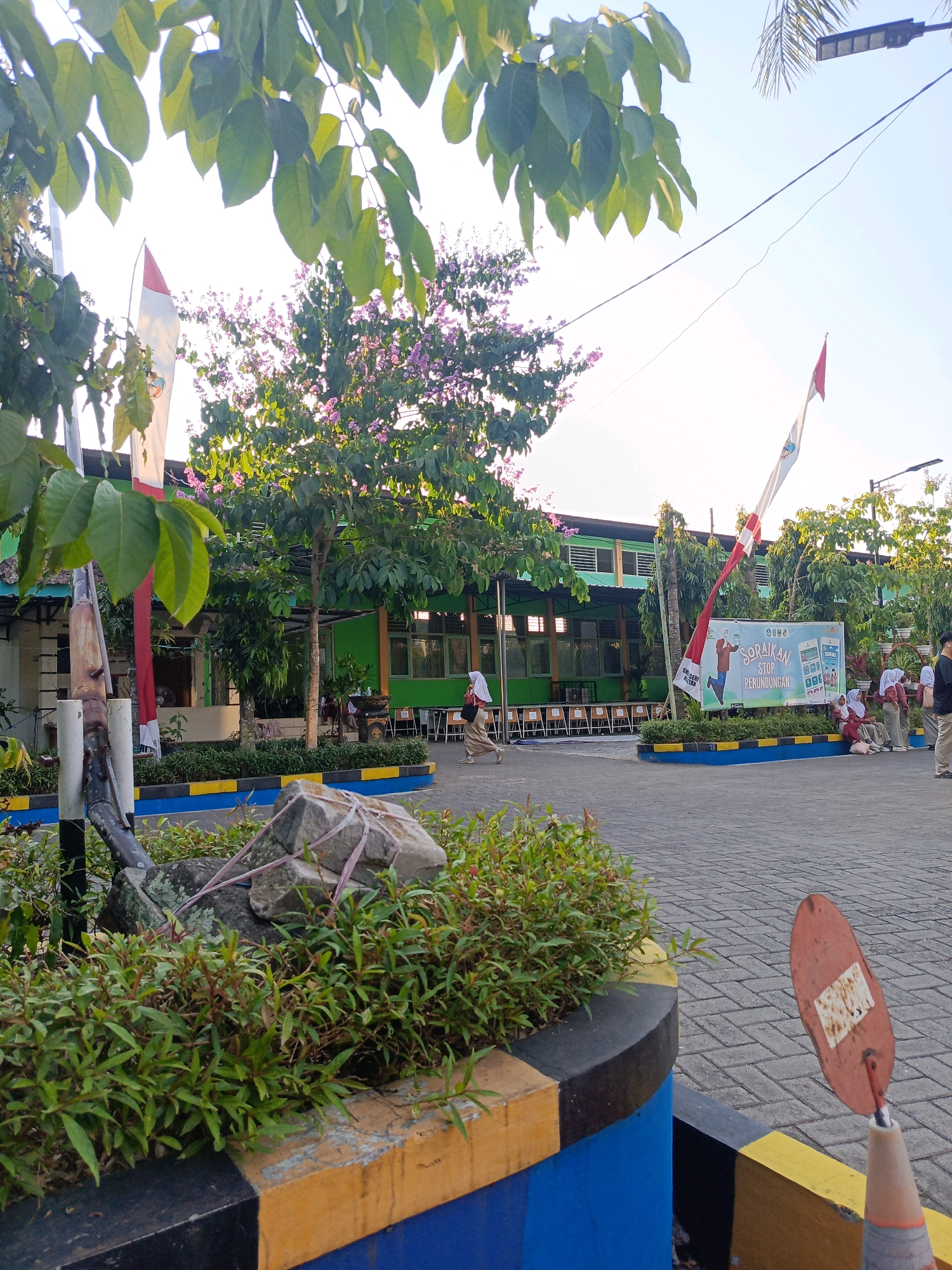 In Candirejo, Indonesia, this outdoor image captures a school courtyard with a vibrant green building in the background. Several students, dressed in school uniforms, are visible. Some are walking, while others are seated on benches or the ground, conversing. A large banner with Indonesian text and illustrations is displayed, promoting a campaign. Two Indonesian flags, red and white, are planted in front of the building. Lush green trees and flowering plants adorn the courtyard, with a well-maintained planter box in the foreground. The ground is paved with interlocking bricks. The sky is clear and bright, indicating a sunny day.
