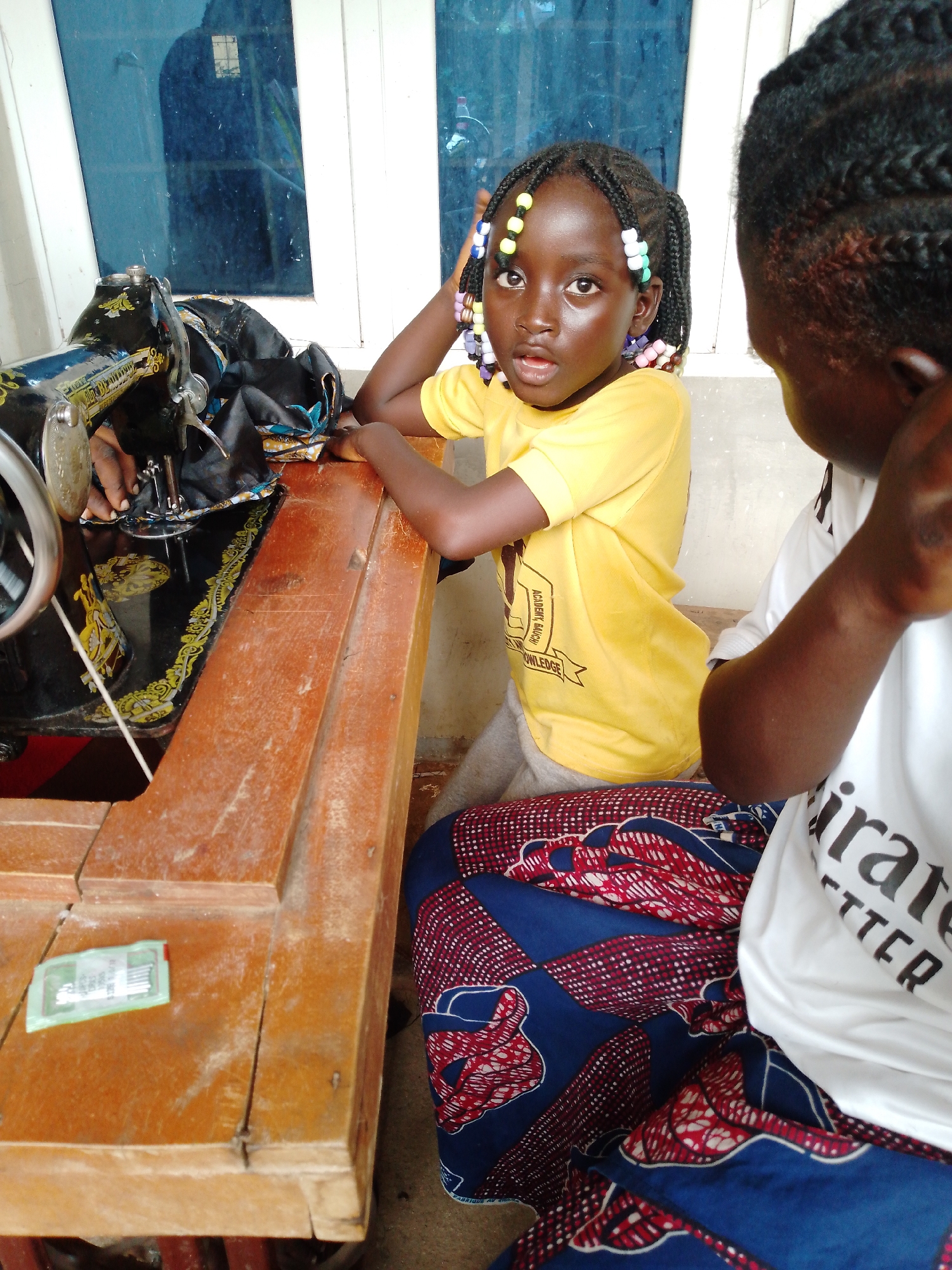 An over-the-shoulder, medium shot captures two young girls near a wooden table with a sewing machine, against a backdrop of blue-tinted windows. In the foreground, a vintage black sewing machine with ornate gold detailing sits on the table. Fabric, black with gold and blue patterns, is draped over the machine and table. A small, green package of sewing needles rests on the table to the left.
The girl on the left, facing forward, has dark skin and is wearing a bright yellow t-shirt with text that appears to be "ACADEMY, NACH... KNOWLEDGE". Her hair is styled in braids adorned with colorful beads. She is looking directly at the camera with a surprised expression.
The girl on the right, facing away from the camera, has dark skin and is wearing a white t-shirt with black text that partially reads "emirates BETTER". She is wearing a blue and red patterned wrap skirt. Her arm is raised, touching her head.
The wooden table appears worn and scratched. The background shows a section of a wall and the blue-tinted windows, which reflect a dim, indoor environment. The lighting is somewhat diffuse, casting subtle shadows. The overall scene suggests a domestic or workshop setting, possibly in a warm climate.