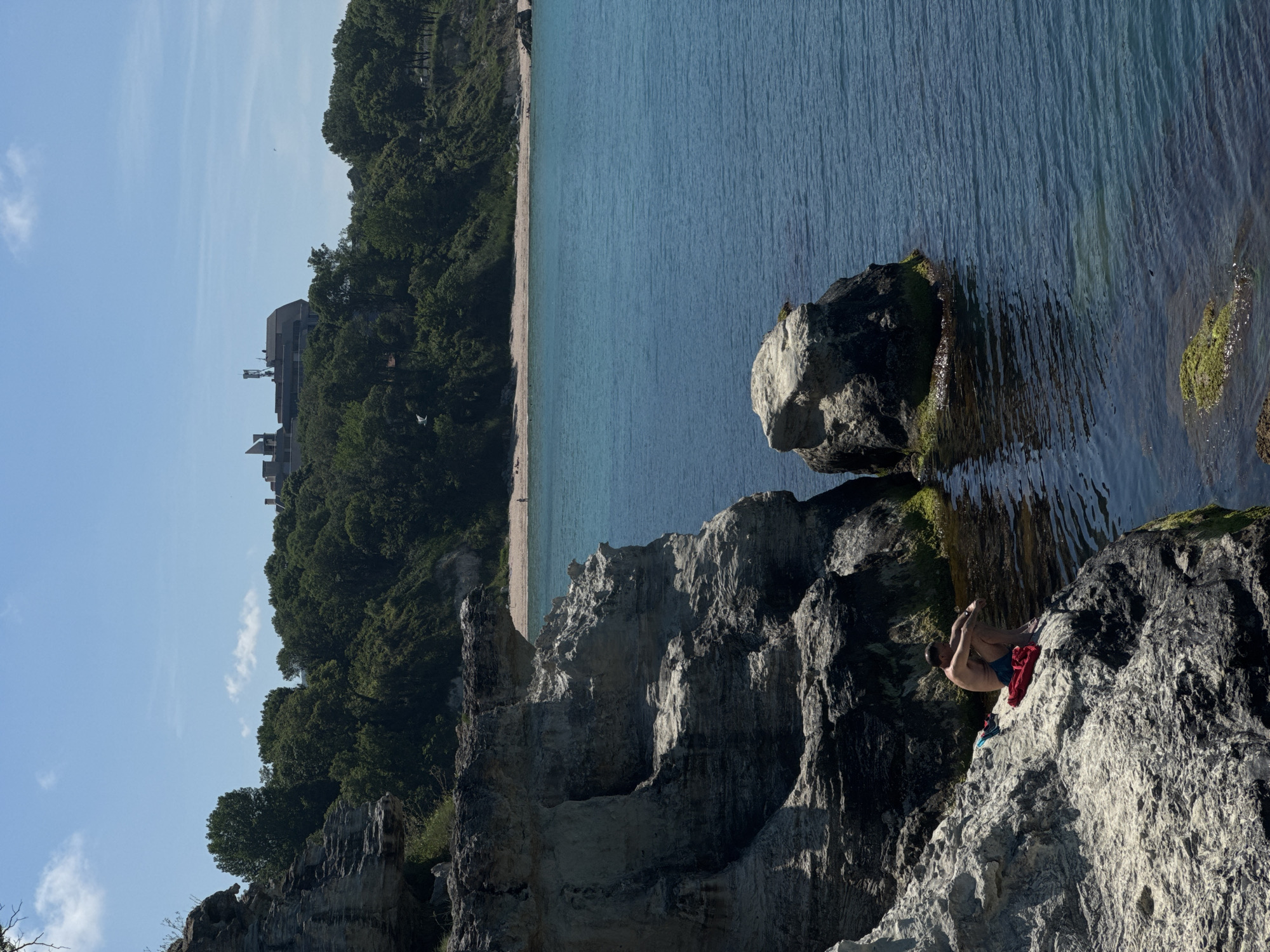 A man sits on a rocky outcrop by the Black Sea in Tsarevo, Bulgaria, with a beach and a building visible in the distance. The man is wearing blue swim trunks and is sitting with his knees drawn up. A red towel and a pair of blue shorts are beside him. The sea is a deep blue, and the sky is clear with a few white clouds. The rocks are gray and weathered, with patches of green moss. The beach is sandy, and there are trees and vegetation on the hillside behind it. A large, modern building with antennas is visible on the hilltop.