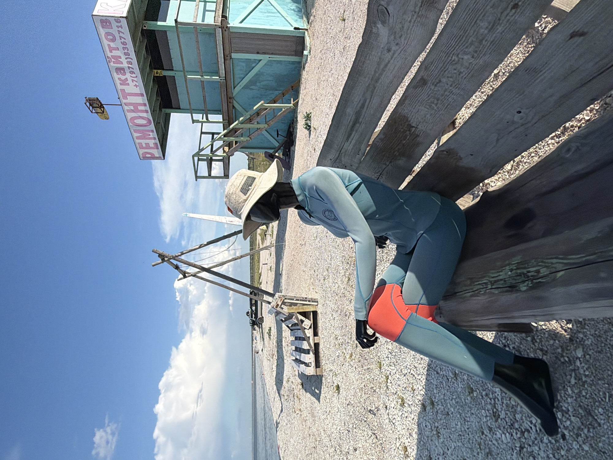 This image depicts a beach scene in Останинское сельское поселение, Ukraine. In the foreground, a mannequin dressed in a teal wetsuit with orange accents and a wide-brimmed hat sits on a weathered wooden bench. The mannequin is positioned to face the left of the frame, with its arms crossed. The beach itself is covered in small white shells.
In the mid-ground, a structure resembling a lifeguard tower or a small building painted teal is visible. It has a sign with pink lettering that appears to say "РЕМОНТ КАЙТОВ" and a phone number. Wooden stairs lead up to this structure. To the left of the tower, a wooden tripod structure stands on the beach, possibly part of a kite or sail rigging. Further back, several vehicles, including what appears to be an SUV, are parked near the water's edge.
The background features a vast expanse of the sea under a bright blue sky with scattered white clouds. The overall impression is of a quiet, possibly seasonal, beach location.