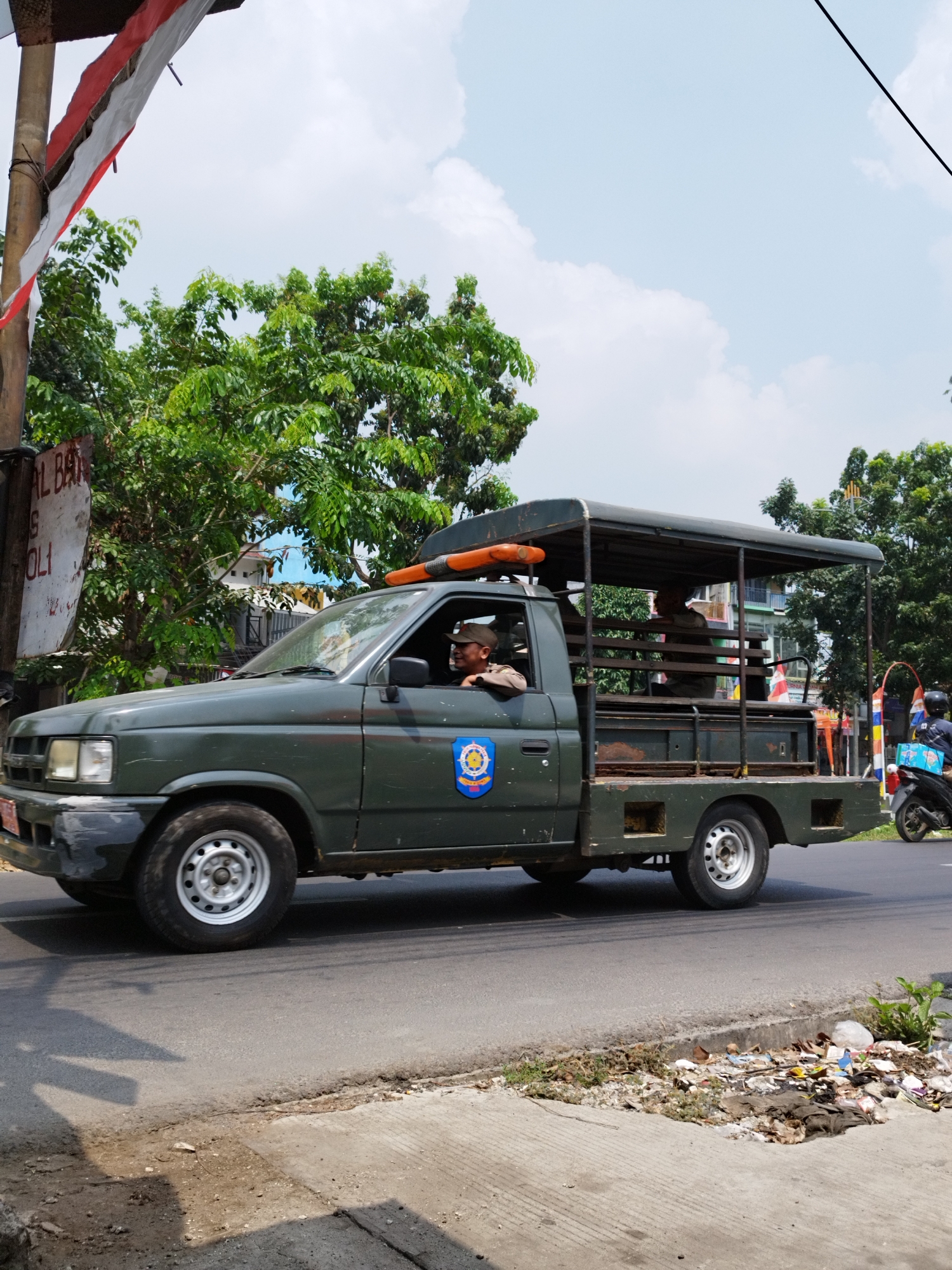A dark green utility truck with a covered bed and a cage-like rear section is traveling on an asphalt road in Bekasi, Indonesia. The truck has two occupants visible. The driver, wearing a cap and a light-colored shirt, is looking forward. Another person, also wearing a cap, is seated in the covered rear of the truck. A blue and yellow emblem is visible on the driver's side door. The truck is moving from left to right in the frame. In the background, there are lush green trees, buildings, and a sky with white clouds. A red and white Indonesian flag is partially visible in the upper left corner, attached to a wooden pole. To the right of the truck, a motorcycle with two occupants is also in motion. The roadside is lined with overgrown grass and debris, suggesting a somewhat unkempt urban environment.