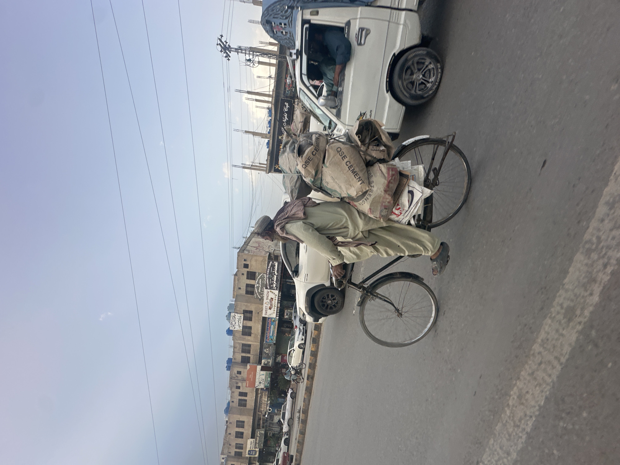 A man is riding a bicycle heavily loaded with bags of cement on a street in Quetta City Tehsil, Pakistan. The bicycle is old and black. The man is wearing traditional clothing and a head covering. He is cycling along a street with parked cars and buildings on the left and other vehicles passing on the right. There are business signs visible in the background, one referencing "Night Cafe" and another listing various shops. Utility poles and overhead wires are visible against the sky.