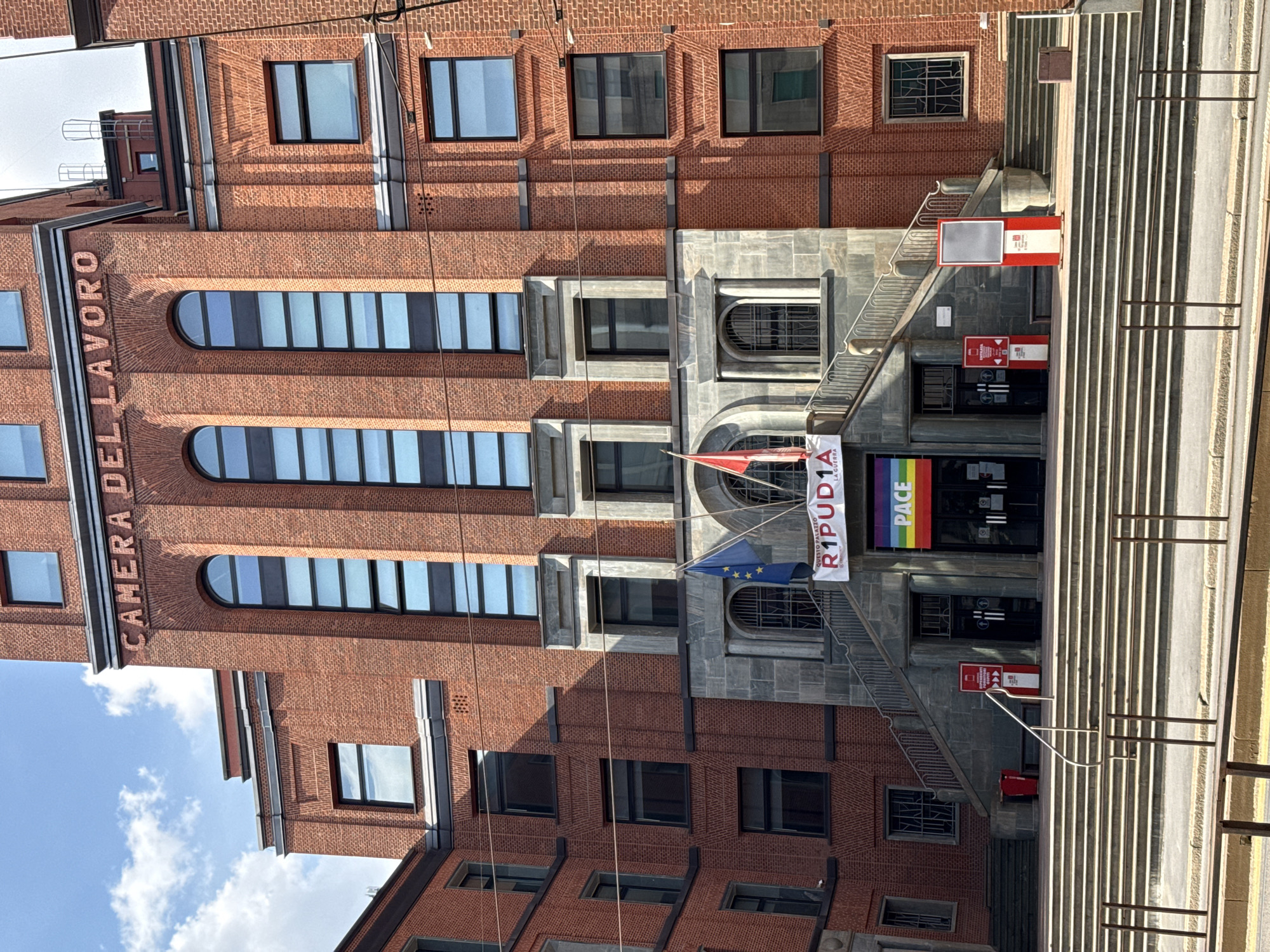 The image depicts the Camera del Lavoro building, located in Milan, Italy. The grand, multi-story structure is constructed primarily of reddish-brown brick with contrasting grey stone architectural elements, particularly around the main entrance and some window frames.
The central facade features a prominent, symmetrical entrance accessed by a wide, double-flight stone staircase that converges at a central landing. Above this landing, three arched doorways, fitted with dark double doors and partially obscured by signage, lead into the building. Two decorative arched windows with iron grates flank these doors.
Above the entrance, a white banner with red text reads "QUESTO PALAZZO R1PUD1A LA GUERRA" (This building REPUDIATES WAR), with additional text "CGIL MILANO" visible. Below this, a horizontal rainbow-striped flag, bearing the white word "PACE" (Peace), is draped across the entrance. To the left of the entrance, an Italian flag (red visible) and a European Union flag (blue with yellow stars) are displayed on poles.
On the ground level, flanking the bottom of the main staircase, two red and white information stands, one displaying "ENTRATA" (Entrance), are present. An additional, similar stand, appearing to be an empty display board, is situated to the right of the staircase's upper landing. A red waste bin is positioned to the left of the staircase.
The upper levels of the central section feature three tall, narrow arched windows with multiple panes, reflecting the sky. Above these windows, the building's name "CAMERA DEL LAVORO" is prominently inscribed in raised red letters. The side wings of the building display multiple rectangular windows with dark frames, varying in size and arrangement across different floors. The facade incorporates recessed brickwork and stone cornices as decorative elements.
Overhead utility wires traverse the mid-ground of the image. The immediate foreground shows a series of lower steps and metal railings, suggesting a public thoroughfare or tram line is just out of frame. The sky above is clear blue with scattered white clouds. No individuals are visible in the scene.