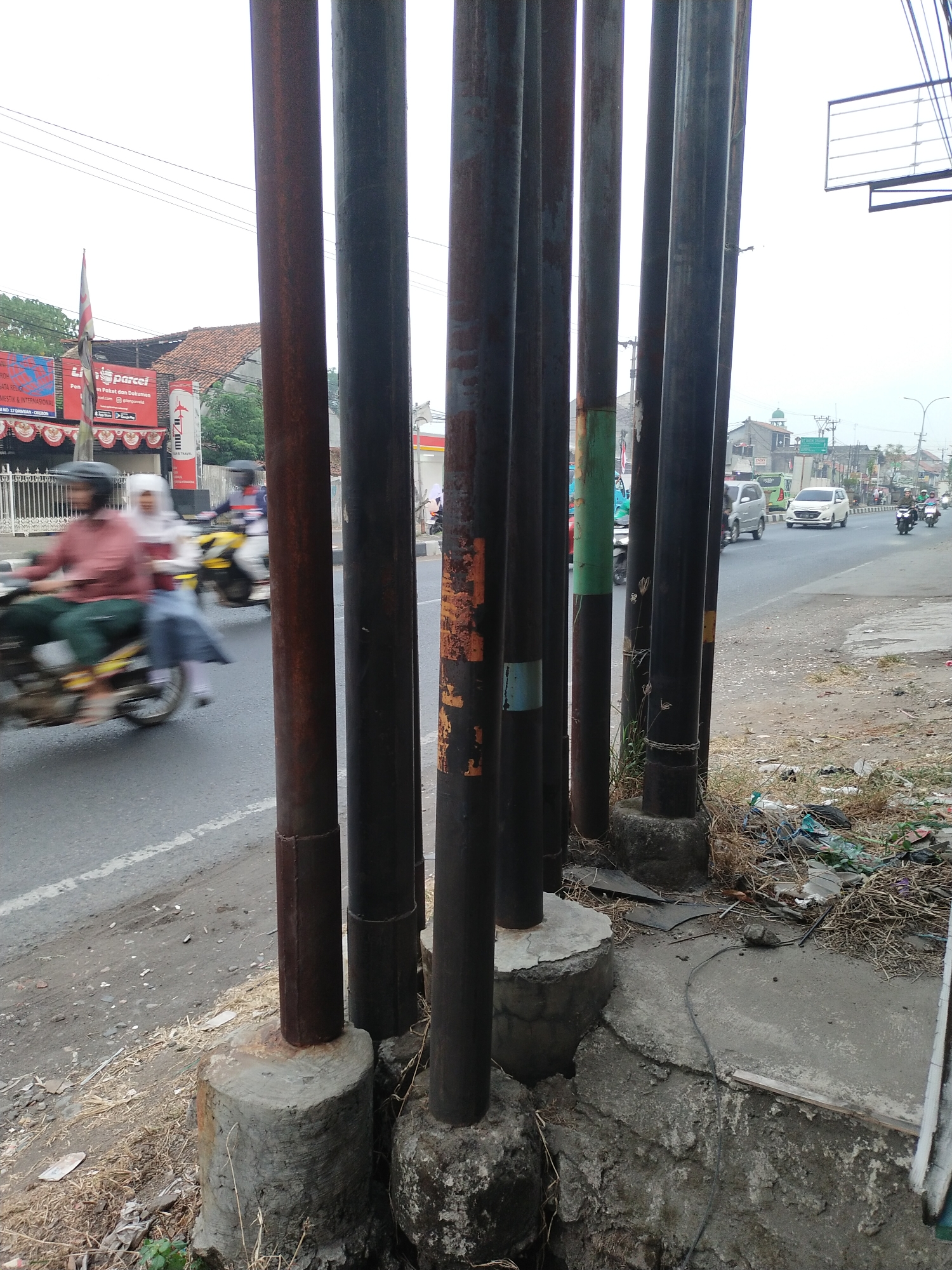 The image depicts a street scene in Kemlakagede, Indonesia, with a prominent line of dark metal poles in the foreground. These poles are weathered and show signs of rust, some with patches of orange and blue paint. They are anchored in concrete bases that are uneven and cracked. To the left of the poles, a road is visible with two motorcycles in motion. The rider of the closest motorcycle is wearing a red shirt and helmet, and a passenger in a white headscarf and blue skirt is seated behind them. The motorcycle is yellow and appears to be moving from left to right. Further down the road, other vehicles, including cars and motorcycles, are present, indicating moderate traffic. Buildings, some with tiled roofs, and trees line the street in the background. A flagpole with a red and white flag is visible on the left side of the image, near a building with red awnings. The ground along the side of the road, next to the poles, is covered with dry grass, debris, and litter, suggesting a less-maintained area. The sky is overcast.