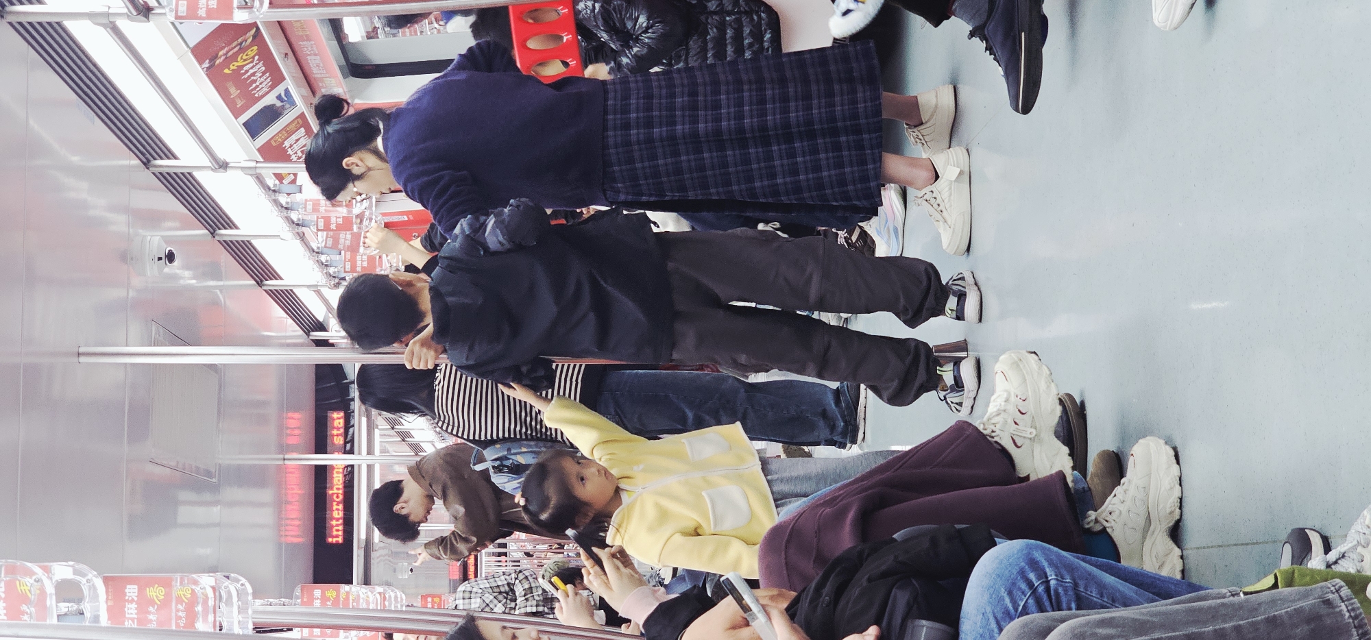 A wide-angle, eye-level shot shows a subway car with several passengers standing and sitting during the daytime in Yuzhong District, China. The subway car has shiny metal walls with red advertisements. Passengers of different ages are dressed in a variety of clothing, including jackets, sweaters, jeans, and skirts. Many are holding onto metal bars. Some passengers are looking down at their phones. The floor is light blue-gray.