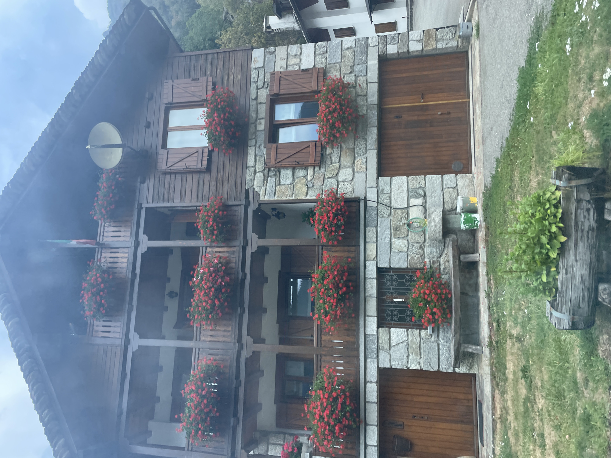 The image shows a multi-story wooden and stone building in Piode, Italy, with a clear blue sky overhead. The building has a dark wooden roof and dark wooden siding on the upper stories, with large stone blocks forming the lower story and parts of the upper stories. There are multiple balconies adorned with abundant red flowers in planters. Several windows are visible, some with wooden shutters. On the ground level, there are two large wooden garage doors and a wooden bench positioned in front of a barred window. In front of the building, there is a grassy area with some white wildflowers and a wooden trough planter in the foreground. The overall scene is picturesque and well-maintained.