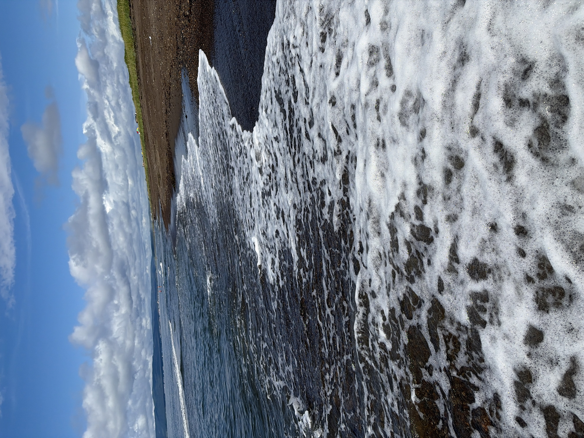 The image depicts a view of the sea shore in Ozerskoe, Russia, under a partly cloudy sky. The foreground is dominated by the dark, wet sand of the beach, with the white foam of incoming waves covering a significant portion of the frame. The sea itself is a deep blue, with visible ripples and waves extending towards the horizon. In the background, a grassy incline rises from the beach, with a few small structures and tents visible at its crest, suggesting a possible campsite or settlement. The overall scene is one of a natural coastal environment under a dynamic sky.