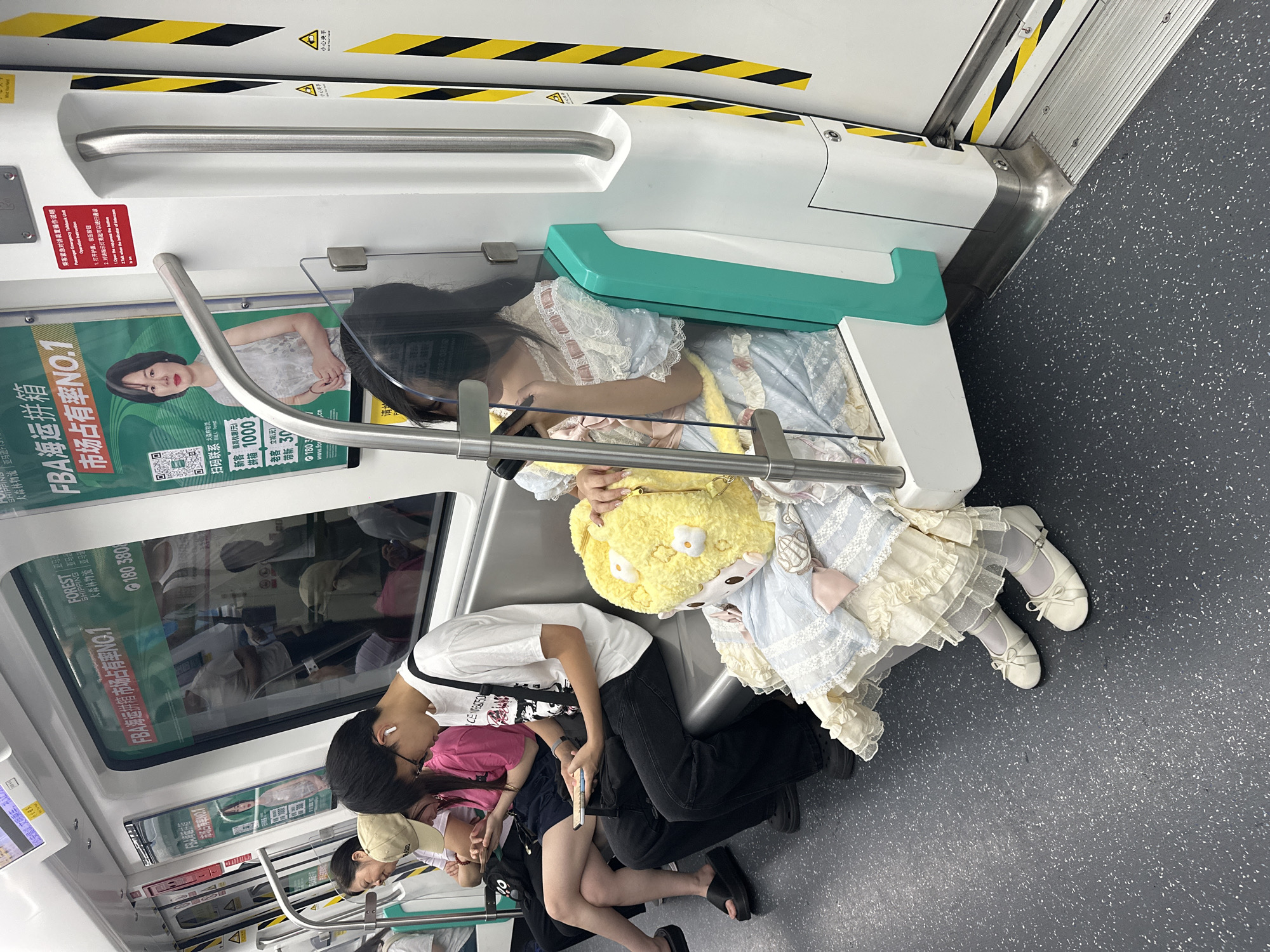 The image shows the interior of a subway car with several passengers seated. In the foreground, a young girl in a light blue and white lace dress and white ballet flats is holding a large yellow plush toy. To her left, a young man in a white t-shirt and black pants is looking down at his phone. Further to the left, a young woman in a pink shirt and dark shorts is also looking at her phone, and behind her, another woman with a tan baseball cap is seated. The subway car has gray seats and a dark gray floor. Advertisements are visible on the windows of the train, reflecting the passengers inside. The overall scene depicts a typical commute on public transportation.
