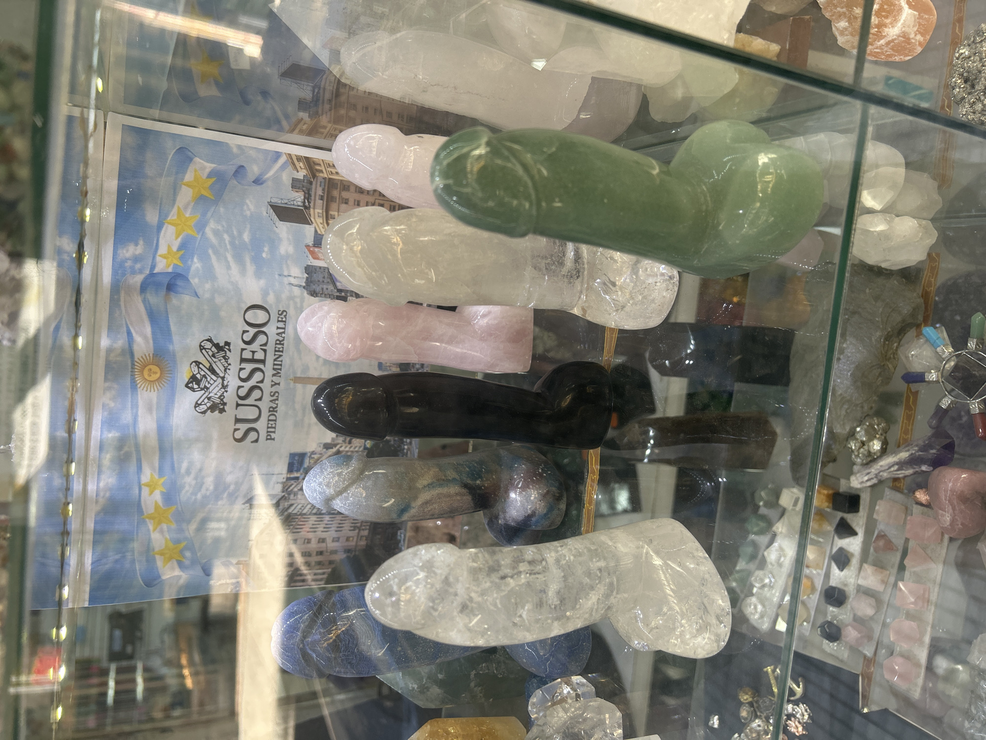 The image depicts a glass display case containing several phallic-shaped objects carved from various minerals. The objects are arranged on top of a promotional advertisement for "SuSSeSo Piedras y Minerales," suggesting the location is a shop in Buenos Aires, Argentina that specializes in stones and minerals. The advertisement includes an image of the Argentine flag and a cityscape. The display case also contains other mineral specimens and jewelry. The environment appears to be a retail space, with reflections visible on the glass surfaces. There are no people visible in the image.