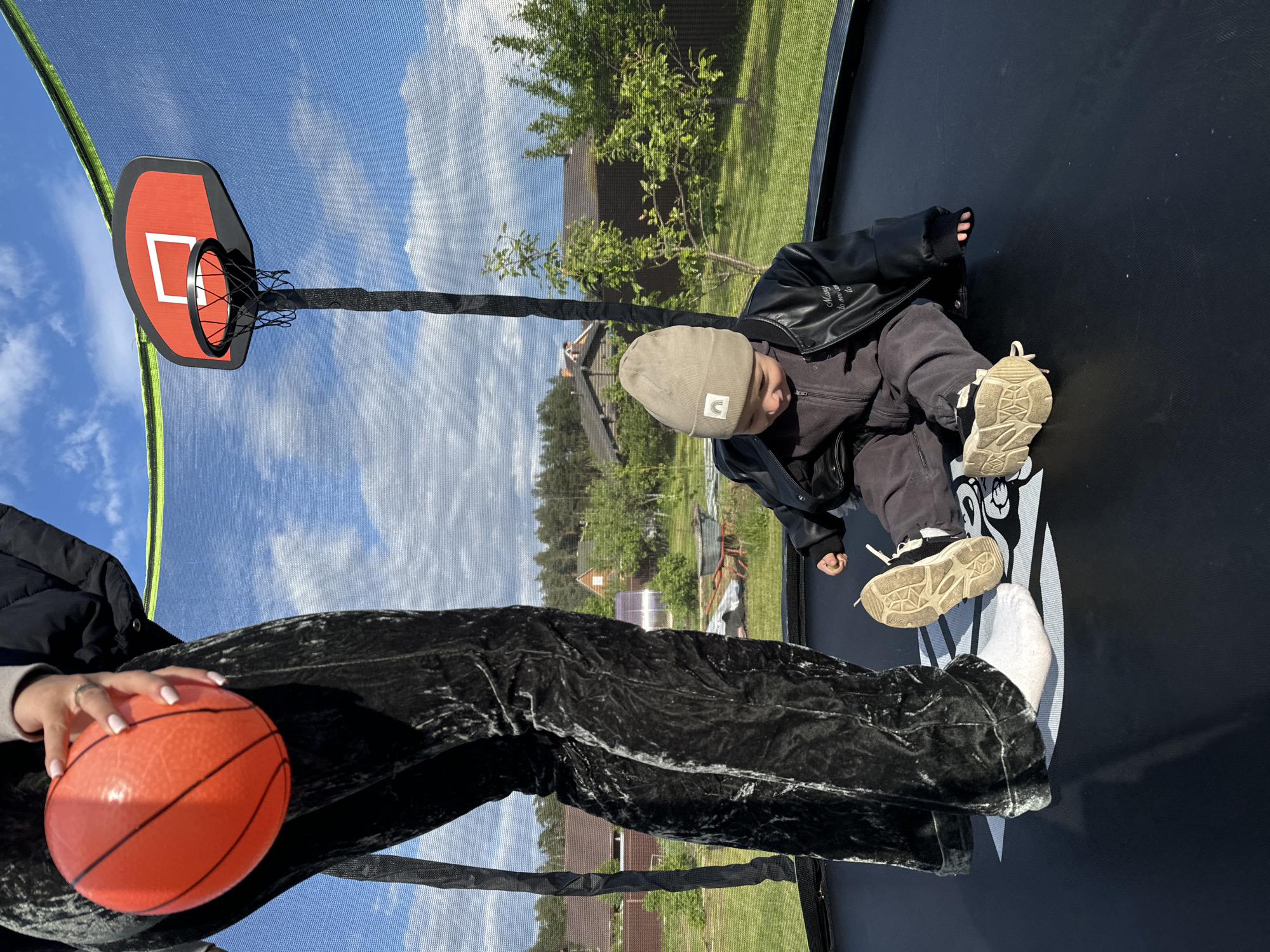 The image depicts a child wearing a beige beanie, dark jacket, and shoes, sitting on a trampoline. Next to the child, a person holds an orange basketball. A basketball hoop with a red backboard is attached to the trampoline net. The setting is outdoors, with trees and buildings visible in the background. The location appears to be Vysokinichi, Russia.