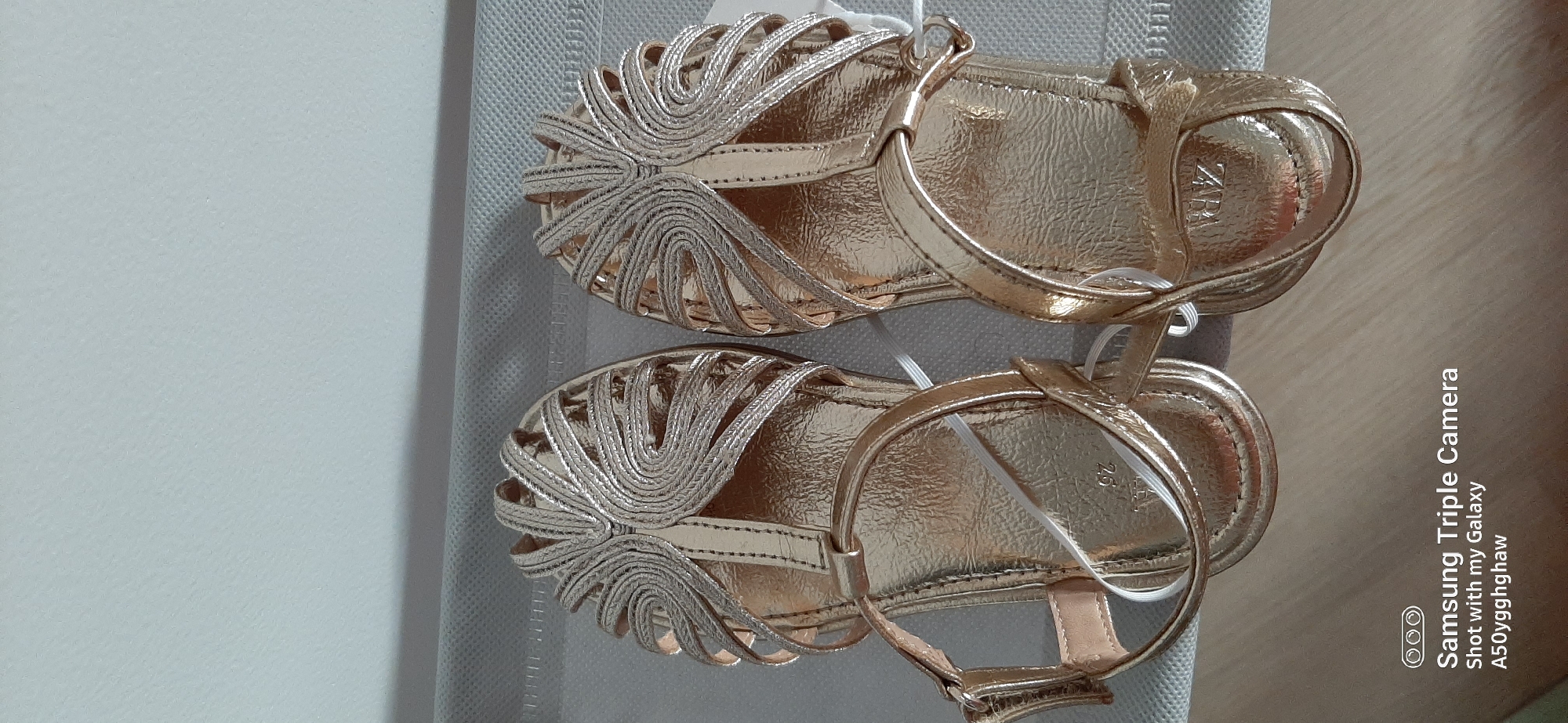 A pair of metallic gold-colored sandals are placed side-by-side on a textured, light gray surface. The sandals have an open-toe design with a decorative strap across the front, resembling a fan or feather motif, made of interwoven thin straps. A thin ankle strap with a buckle closure is attached to the side of each sandal. White elastic straps are visible within the sandal straps. The brand name "ZARA" is embossed on the insole of the right sandal, and a size "25" is visible on the insole of the left sandal. The background is a plain, light-colored wall. The bottom right corner of the image contains a watermark with "Samsung Triple Camera" and "Shot with my Galaxy A50yqghghaw". There is no discernible information to suggest the location is in Dubai, United Arab Emirates, as the image contains no landmarks, signs, or any other contextual clues.