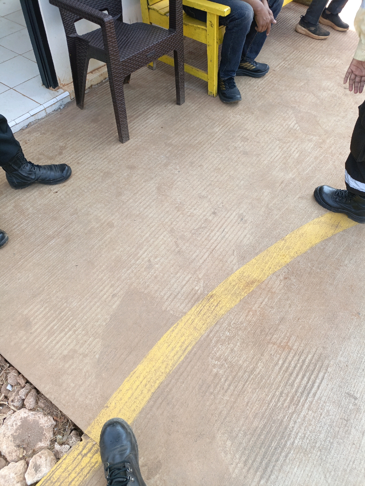 The image shows a group of people's feet and lower legs standing on a textured, light brown concrete surface marked with a yellow line. The scene appears to be outdoors, possibly in a public area. A dark brown woven plastic chair is visible to the left, as is a sliver of a white tiled wall. To the right, the legs of a person in a yellow garment are partially visible. Several individuals are wearing dark-colored boots or shoes. One person is wearing black work boots. Another person is wearing dark sneakers with beige soles. The concrete surface has parallel lines scored into it, suggesting it might be a sidewalk or pathway. The overall impression is of a group of people waiting or gathered in an urban or semi-urban environment. No specific landmarks in Jenu, Indonesia, are identifiable from the image itself.
