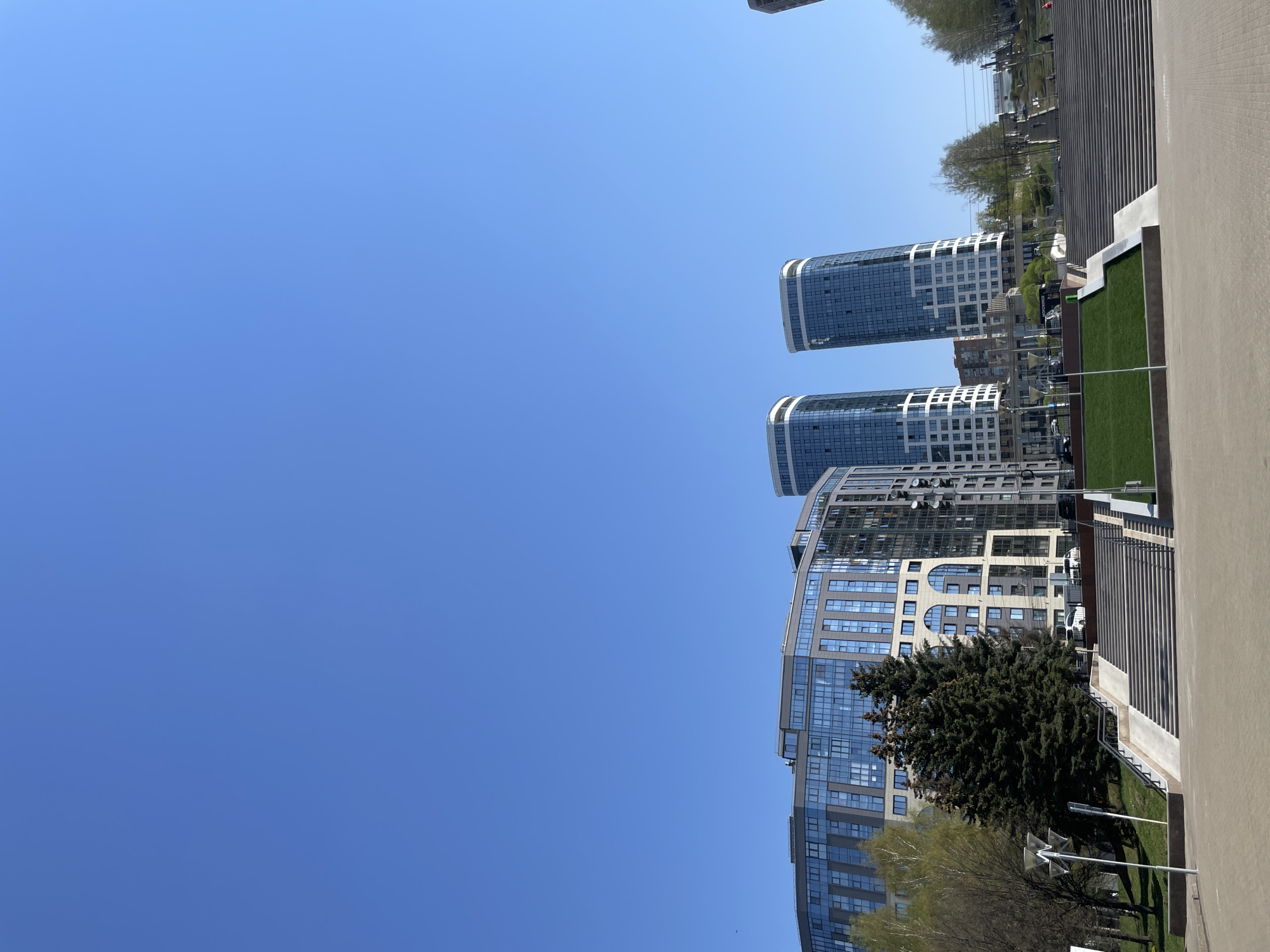 The image shows a street scene in Izhevsk, Russia, with a clear blue sky above. On the right side of the image, there are several modern buildings, including two tall, cylindrical structures with glass facades and white accents. To the right of the buildings, there is a staircase leading to a raised walkway or terrace. The walkway is paved with grey brick. There is greenery visible as well as trees near the base of the buildings. The photo is oriented vertically with the sky dominating much of the frame.